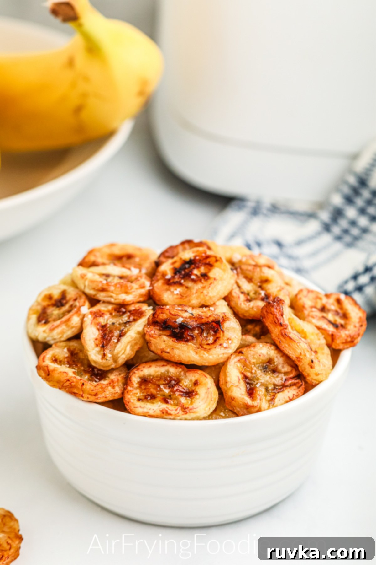 Crispy Air Fryer Banana Chips 3 Banana chips that weere made in the air fryer, served in a white bowl and topped with kosher salt.