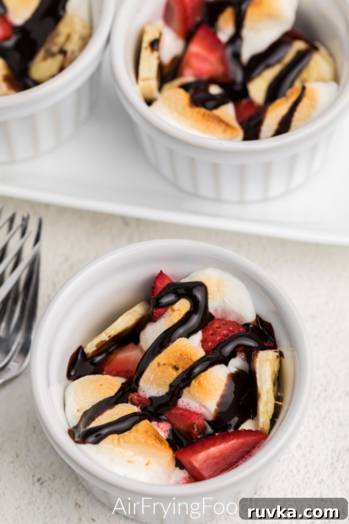 Delicious Air Fryer Strawberry Banana Bowl 2 Ramekin bowls with air fried strawberries, banana, and marshmallows and drizzled with chocolate topping.