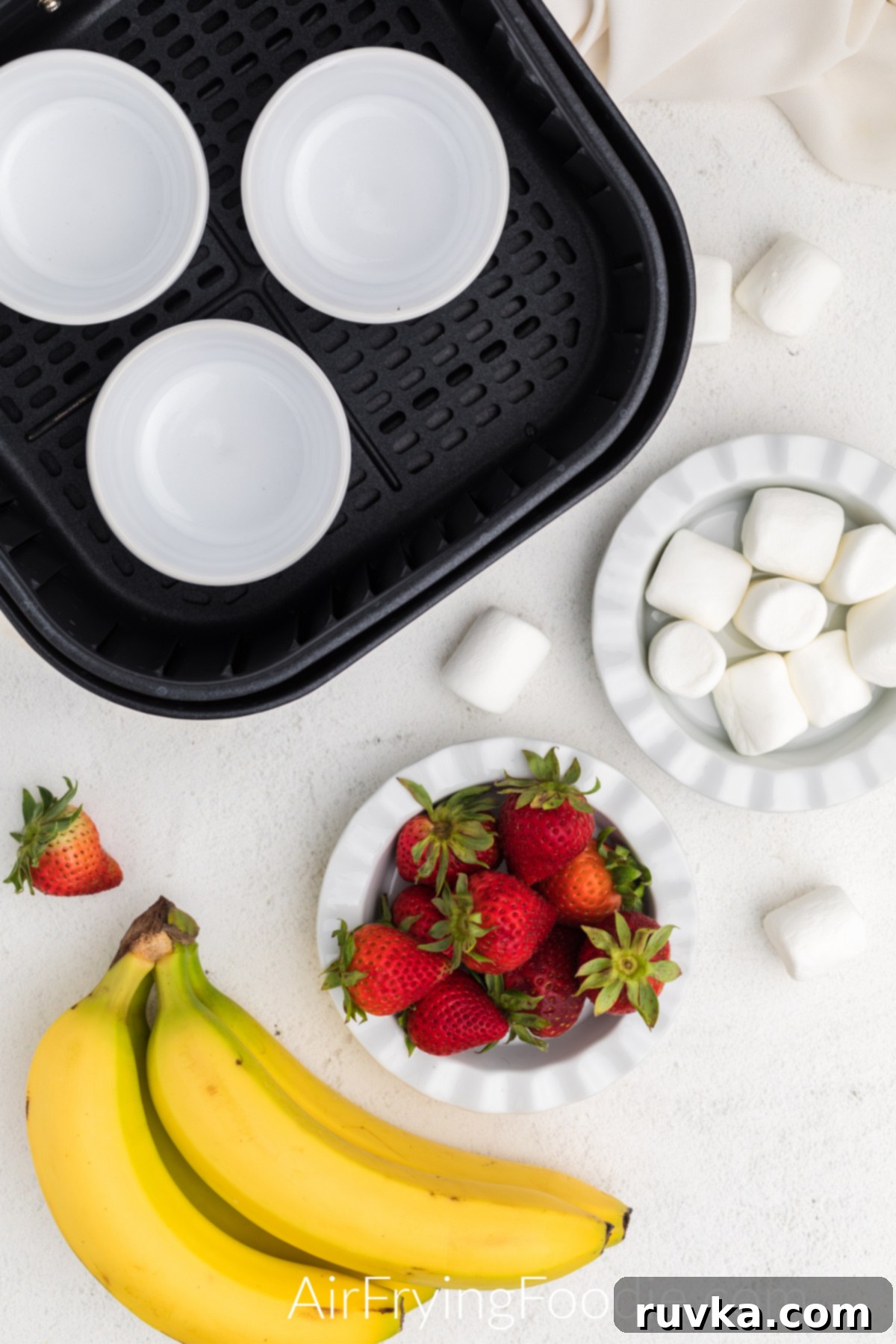 Delicious Air Fryer Strawberry Banana Bowl 3 Air fryer basket with ramekins on a table with bananas, strawberries, and marshmallows, ready for assembly.