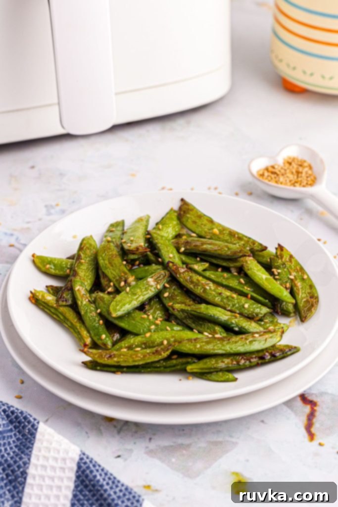 Crispy Air-Fried Snap Peas 2 White plate with crispy air-fried snap peas in front of an air fryer, with sesame seeds scattered on the table for garnish.