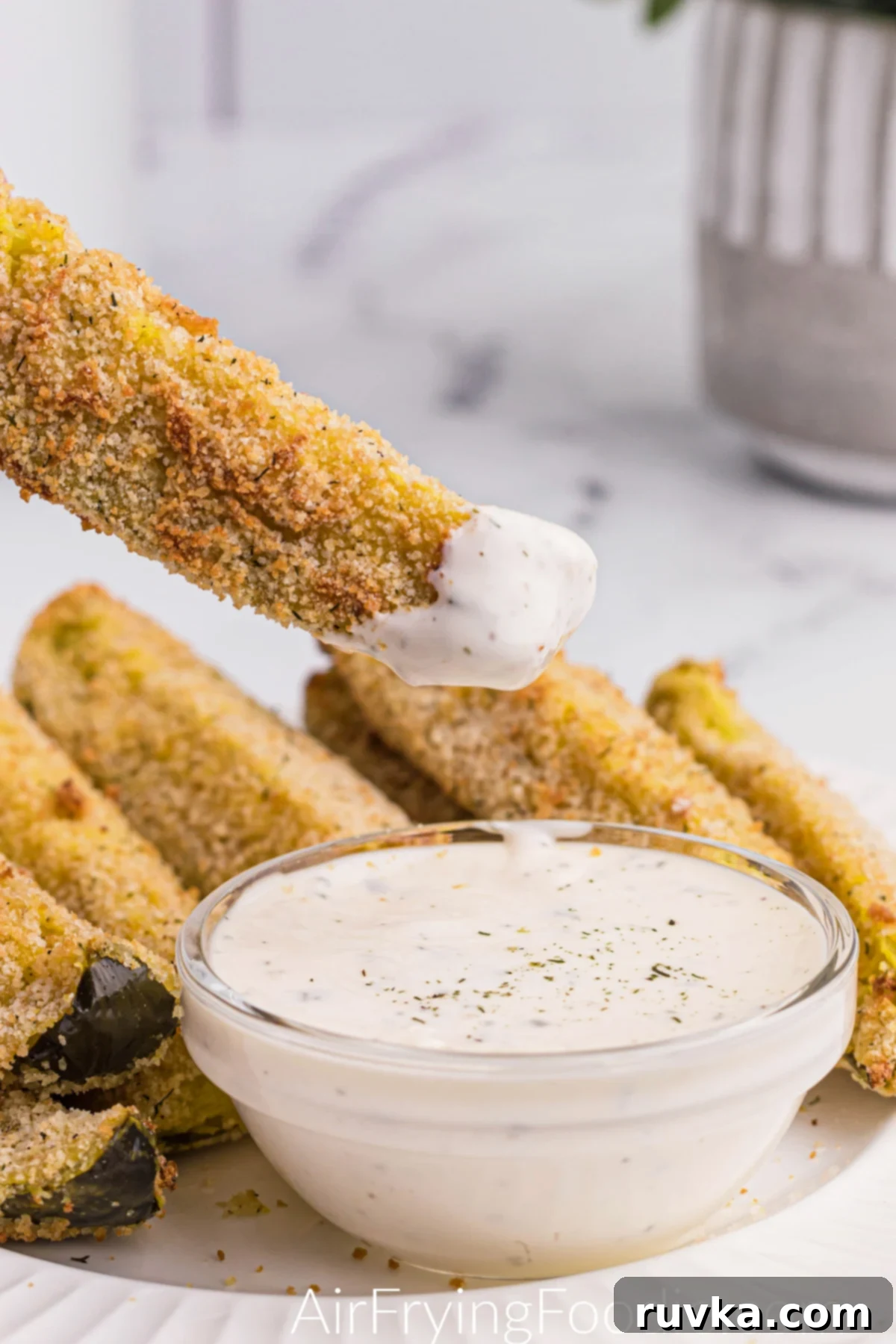 Crispy Air-Fried Dill Pickles 3 Seasoned and breaded air fryer dill pickles on a white plate, with a pickle spear dipped into sauce and readyt o eat.