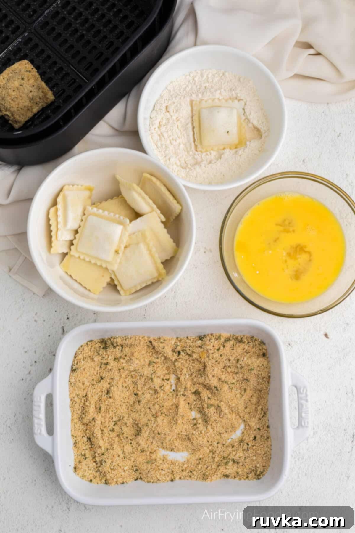 Golden Air-Fried Ravioli Bites 6 Ravioli in bowl of flour after being dipped, and a ravioli in the air fryer basket after being dipped completely.