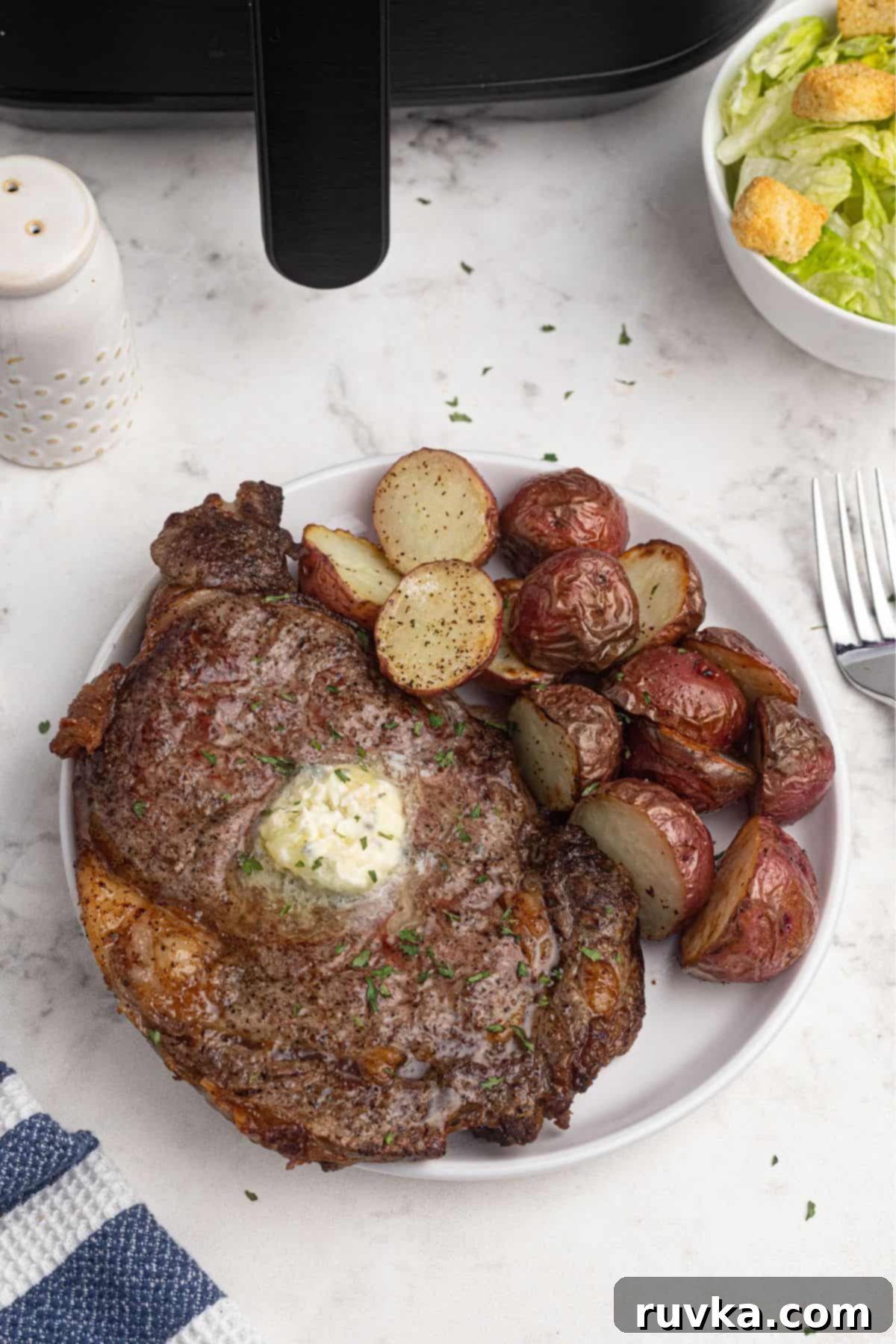 Ultimate Air Fryer Ribeye 2 Juicy air fryer ribeye steak, topped with a pat of butter, on a white plate with baby potatoes.