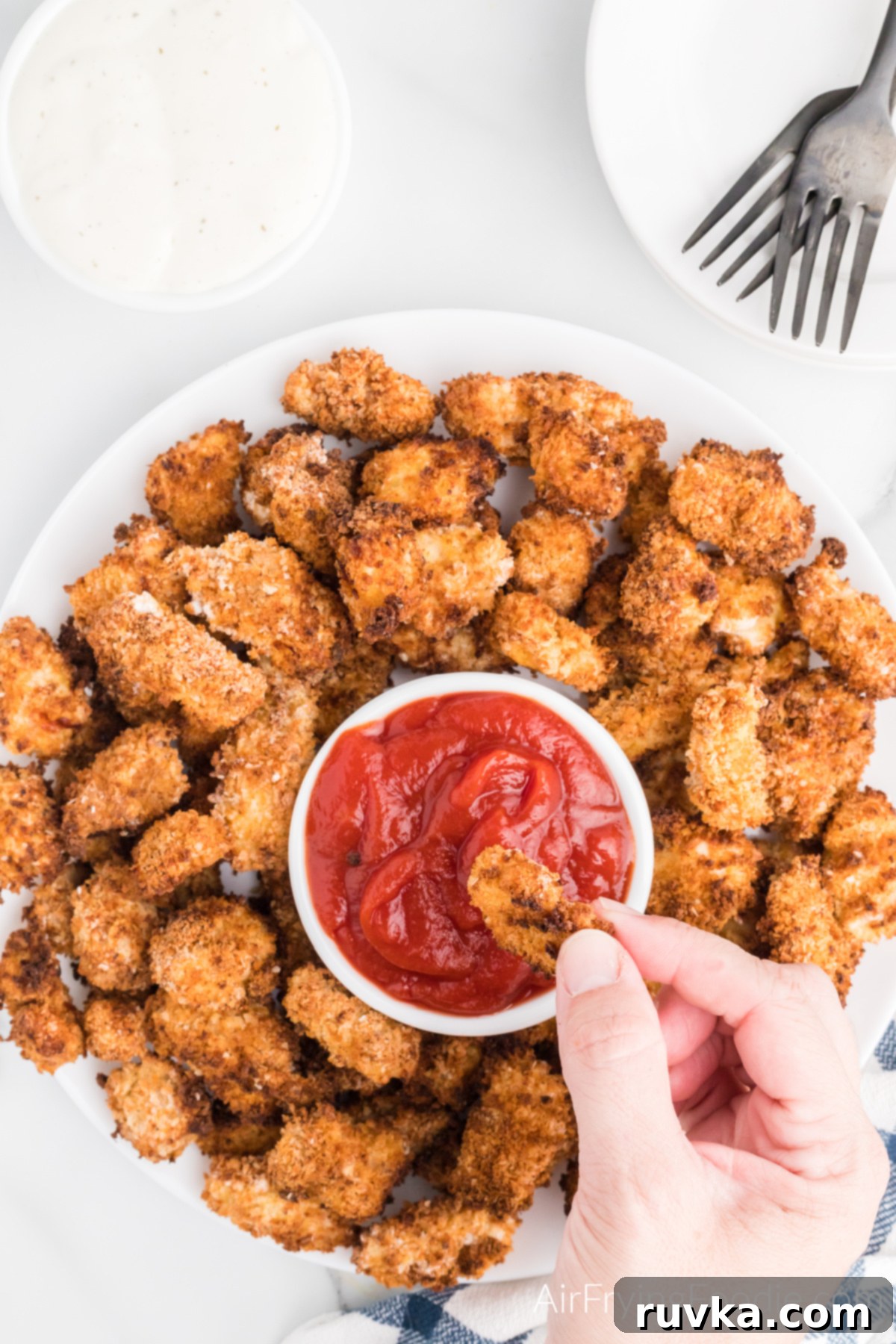 Air-Fried Chicken Poppers 9 Air Fryer Popcorn chicken on a serving plate, with a hand dipping a piece of chicken into a bowl of ketchup.