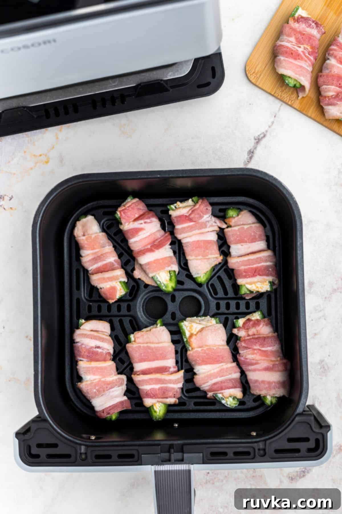 Crispy Air Fryer Jalapeño Poppers 8 Uncooked bacon wrapped jalapenos in the air fryer basket, in front of the air fryer.