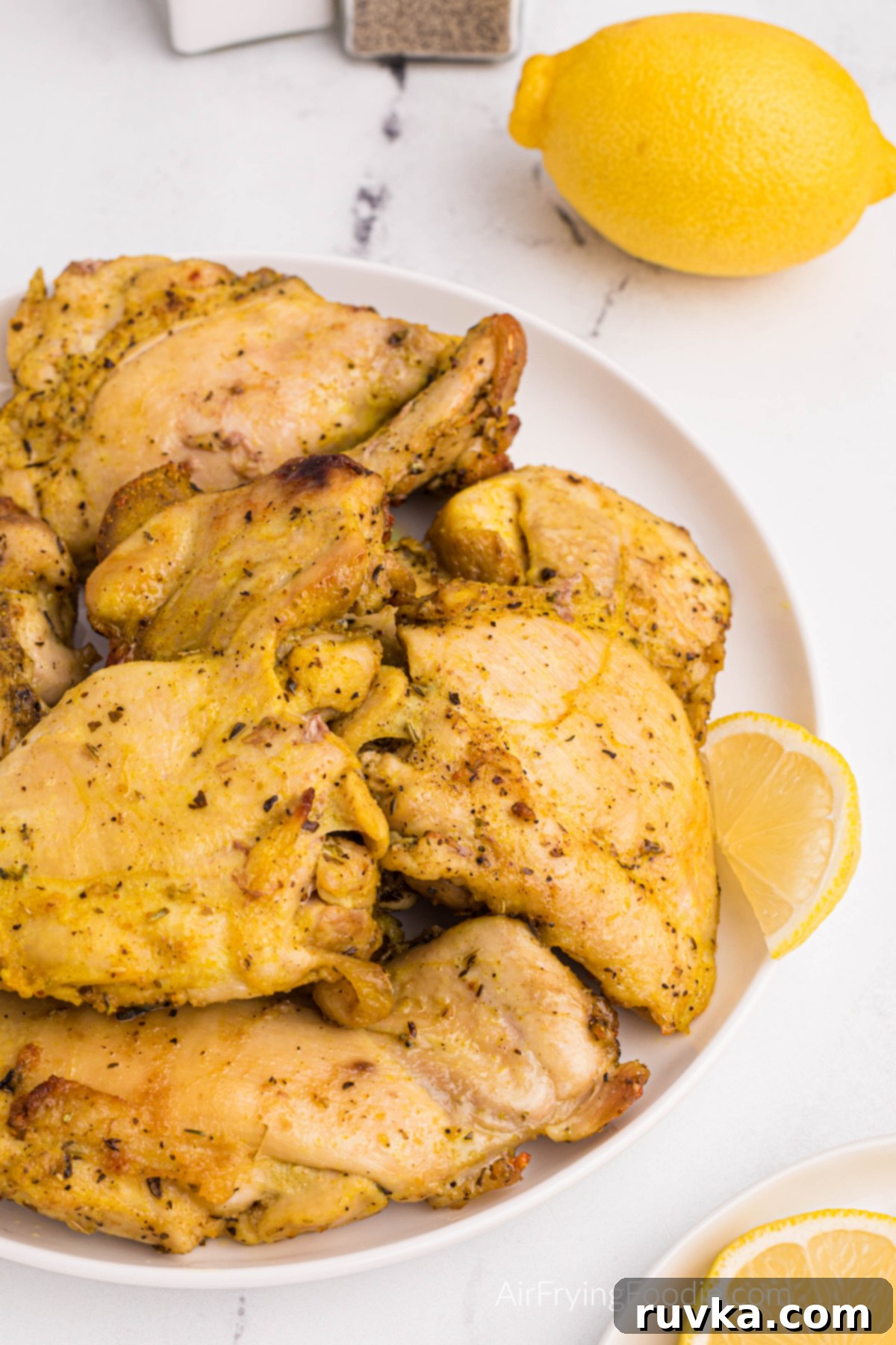 Crispy Air Fryer Lemon Pepper Chicken Thighs 9 Close-up of golden-brown lemon pepper chicken thighs garnished with lemon and fresh herbs on a white plate.
