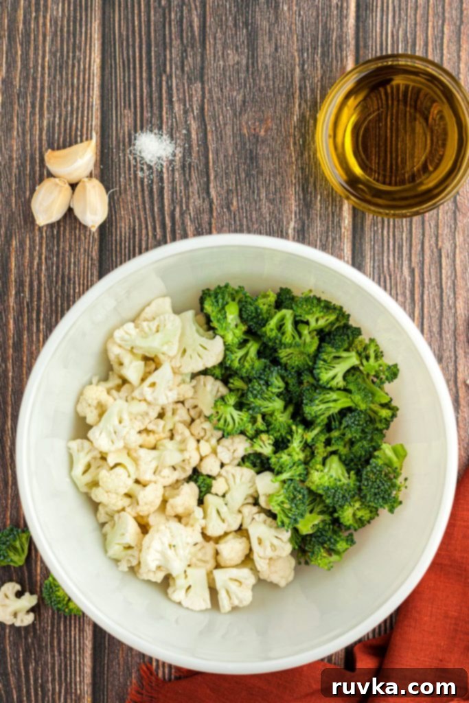 Crispy Air Fried Broccoli Cauliflower 3 Broccoli and cauliflower florets in a white bowl with garlic cloves, salt, and olive oil.