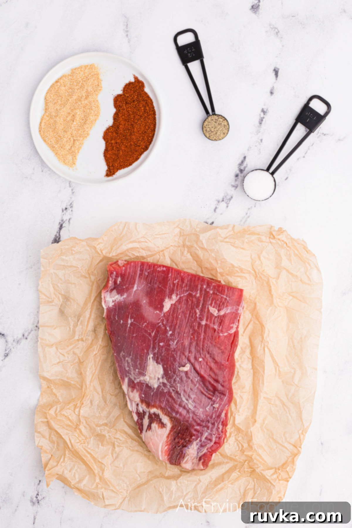 Perfect Air Fryer Flank Steak 3 Flank steak on butcher paper and seasonings in a bowl and in measuring spoons on a white table.