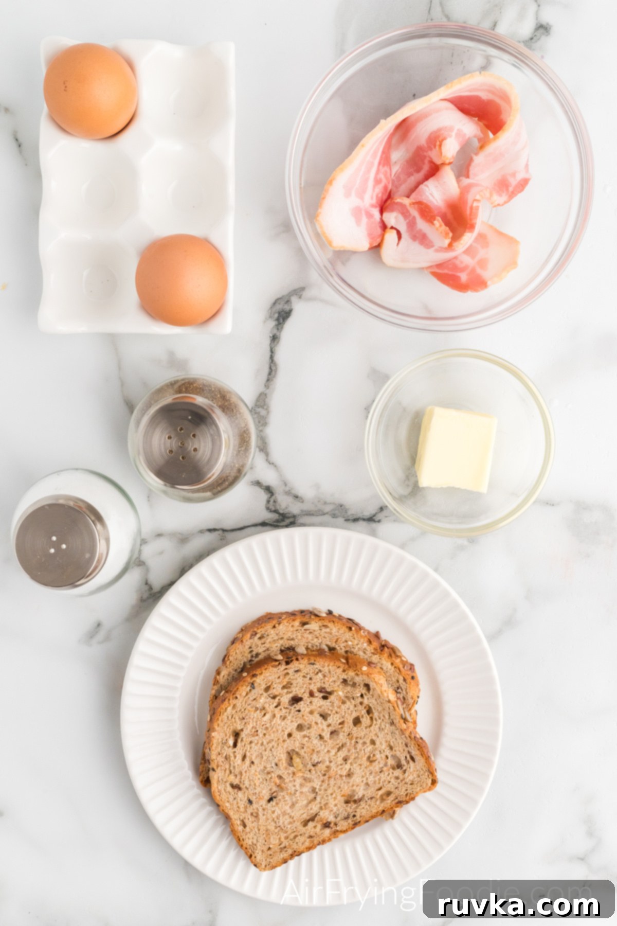 Easy Air Fryer Breakfast Sandwich 3 Overhead view of two pieces of toast on a small round plate, a clear ramekin with butter, a clear bowl with two pieces of bacon, two eggs in a white container, salt, and pepper.