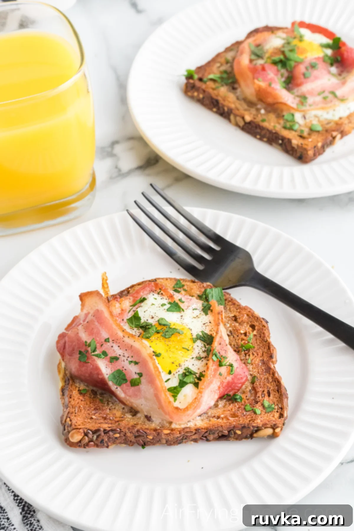 Easy Air Fryer Breakfast Sandwich 9 Fully cooked air fryer breakfast sandwich with bacon and eggs on two round plates sprinkled with parsley served with a glass of orange juice and a fork.