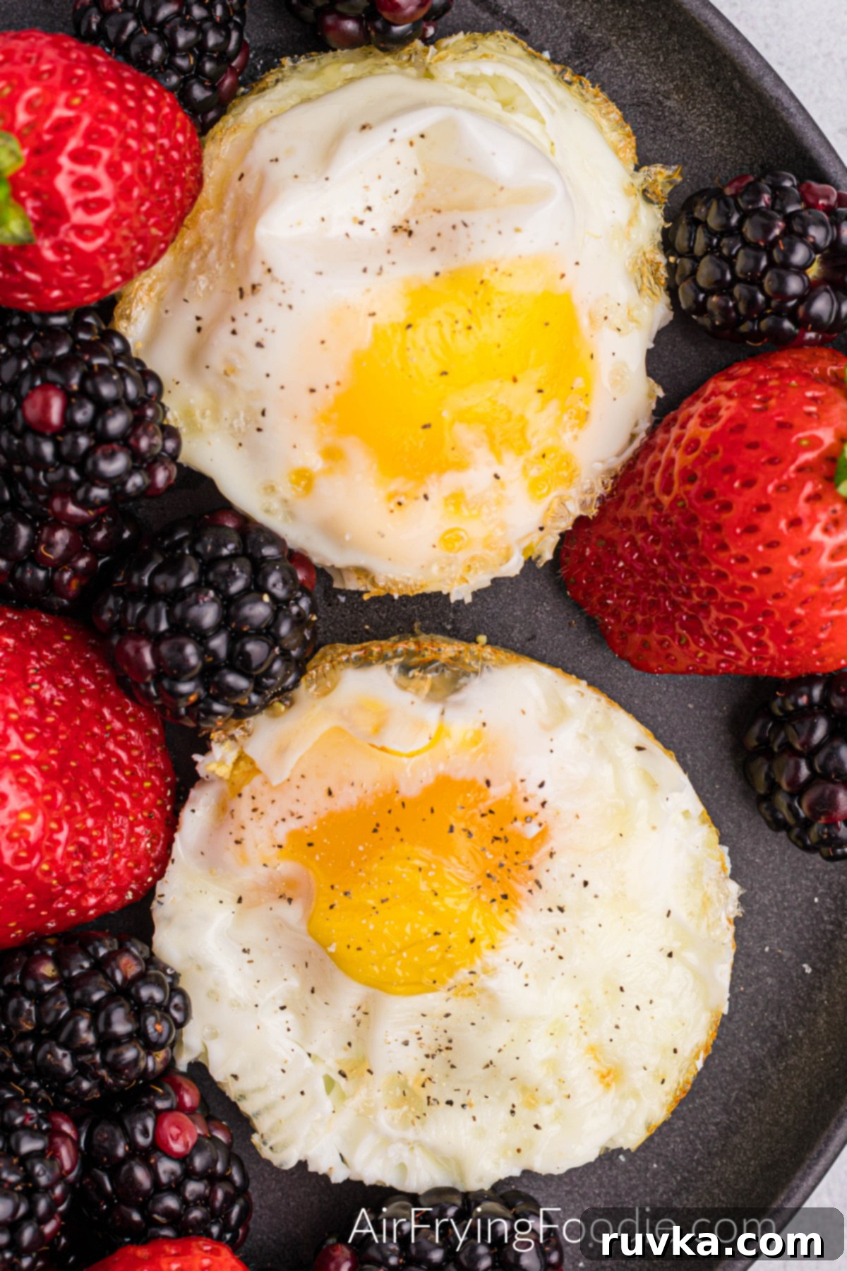 Perfect Air Fried Eggs 7 A close-up of two air fryer fried eggs with a runny yolk, seasoned with salt and pepper, served on a black plate with a garnish of fresh fruit.