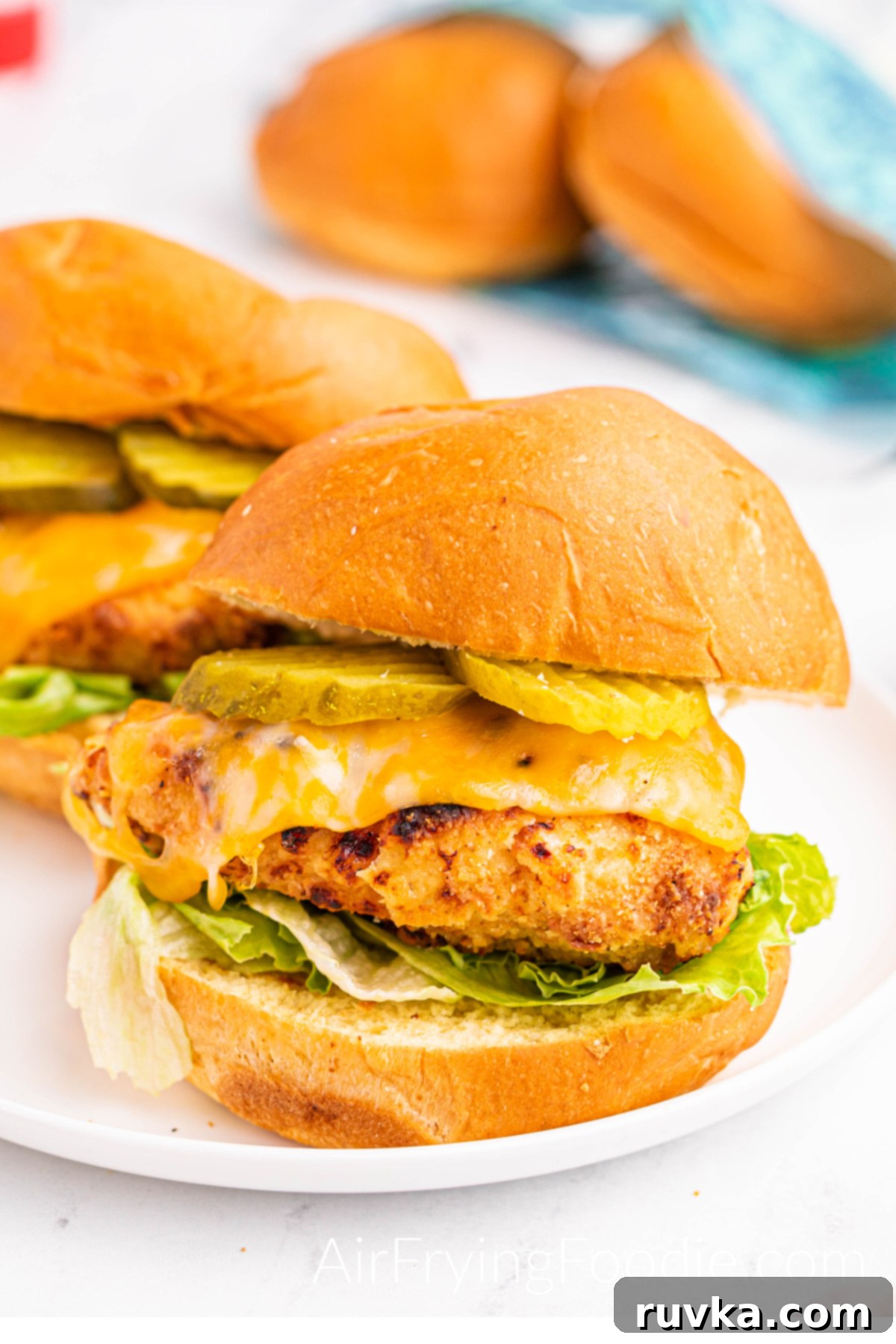 Crispy Homemade Air Fryer Chick-fil-A 2 Two air fryer copycat chick oil a sandwich fully cooked and served on a white round plate dressed with pickles, cheese, chicken breast, lettuce, and chick oil sauce. Two extra buns halfway out of the bag are in the top right of the picture.