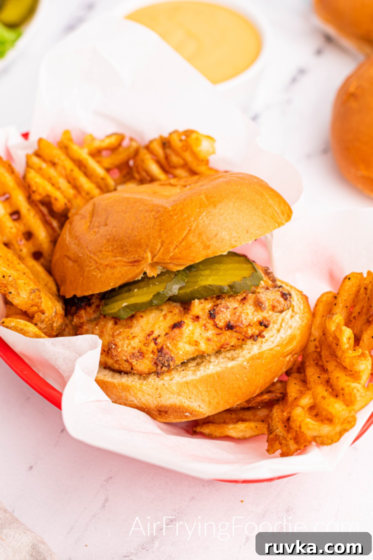Crispy Homemade Air Fryer Chick-fil-A 12 Chicken sandwich fully cooked from the air fryer served with waffle fries in a red basket. There is a white bowl of sauce above the basket and a small portion of extra buns in the image's top right.