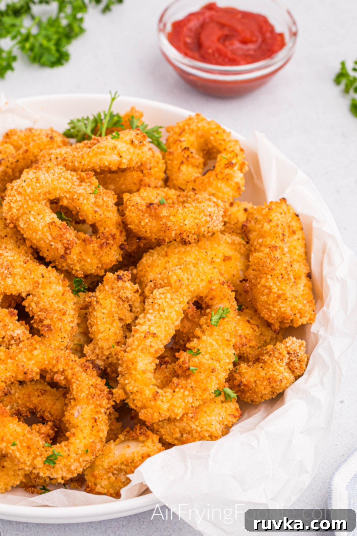 Crispy Air Fried Calamari 2 Fully cooked crispy golden Calamari cooked in the air fry with parsley flakes and served on a round white plate with a side of cocktail sauce in a small clear glass bowl.