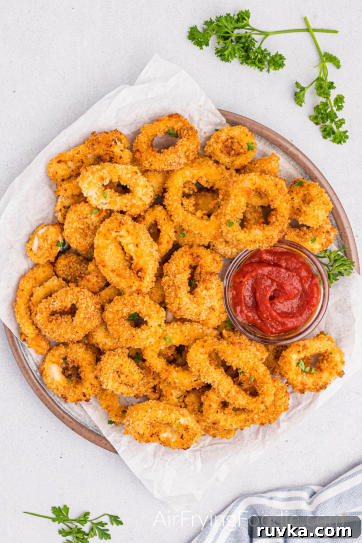 Crispy Air Fried Calamari 11 A tan round plate with crispy calamari fully cooked from the air fryer with a small glass bowl of cocktail sauce with parsley flakes on the calamari and also on the table for looks.