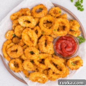 Crispy Air Fried Calamari 13 Close-up of golden brown calamari cooked in the air fryer served on a round white plate with a small glass bowl of cocktail sauce on the plate to the right.