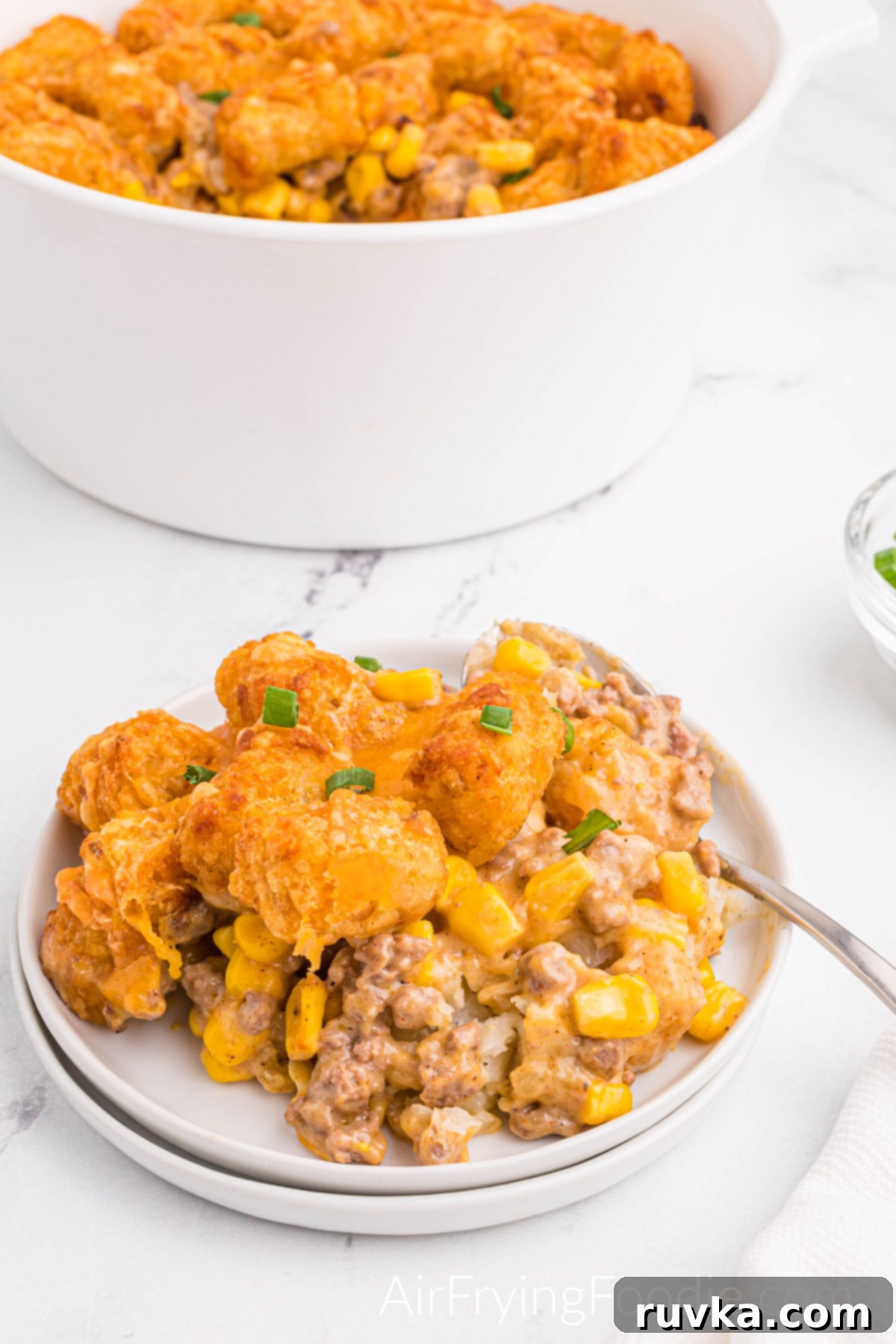Hearty Air Fryer Cowboy Casserole 12 Two small white round plates stacked on top of each other with a serving of cowboy casserole made in the air fryer on the top plate with a fork. The large round white bowl is at the top of the image, with a spoonful of cowboy casserole missing.