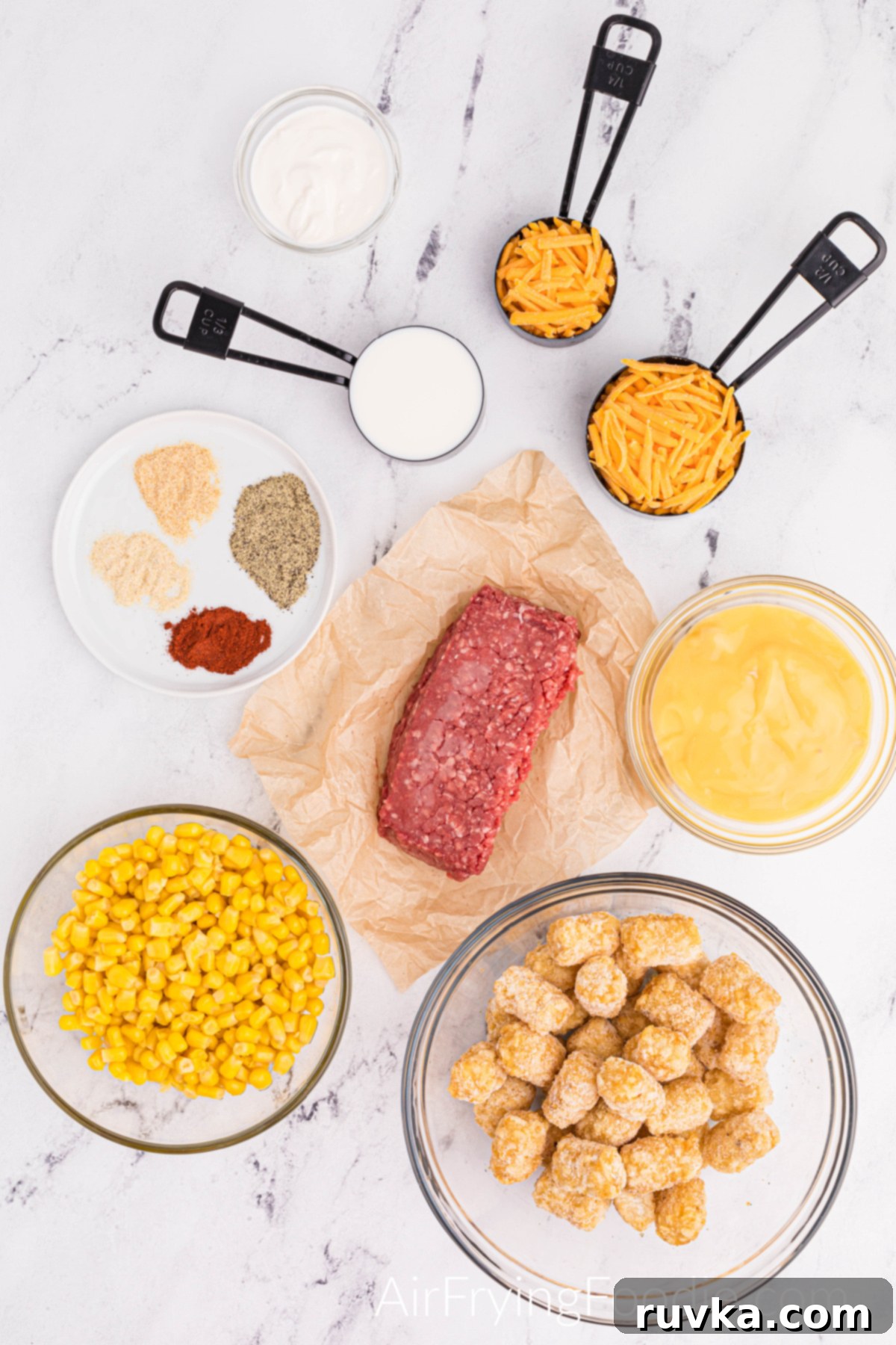 Hearty Air Fryer Cowboy Casserole 3 Ingredients shot with everything you need for cowboy casserole. Four clear bowls contain different elements: corn, tater tots, cream of chicken soup, and sour cream. Raw ground beef in the center of all the ingredients, a round white plate with onion powder, garlic powder, paprika, and ground black pepper. Three measuring cups are measuring; one consists of milk, and the other two measuring cups consist of shredded cheddar cheese.