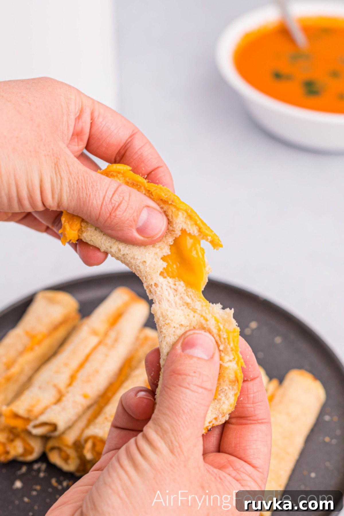 Crispy Cheesy Air Fryer Roll Ups 10 Grilled cheese roll up being torn in half with a black plate filled with grilled cheese roll ups and a cup of tomato soup.