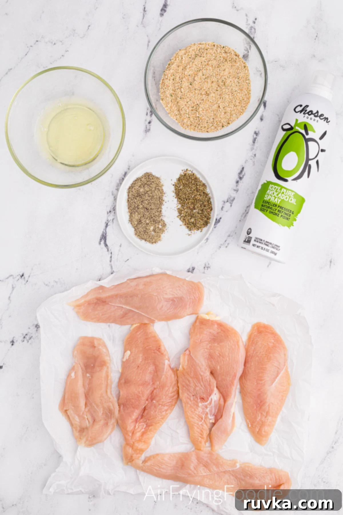 Perfectly Golden Air Fryer Chicken Cutlets 3 Ingredients picture with six boneless, skinless raw chicken breasts, a round white plate with Italian seasoning, and ground black pepper. Two clear bowls consist of egg whites, and the other contains seasoned breadcrumbs and a bottle of olive oil spray.