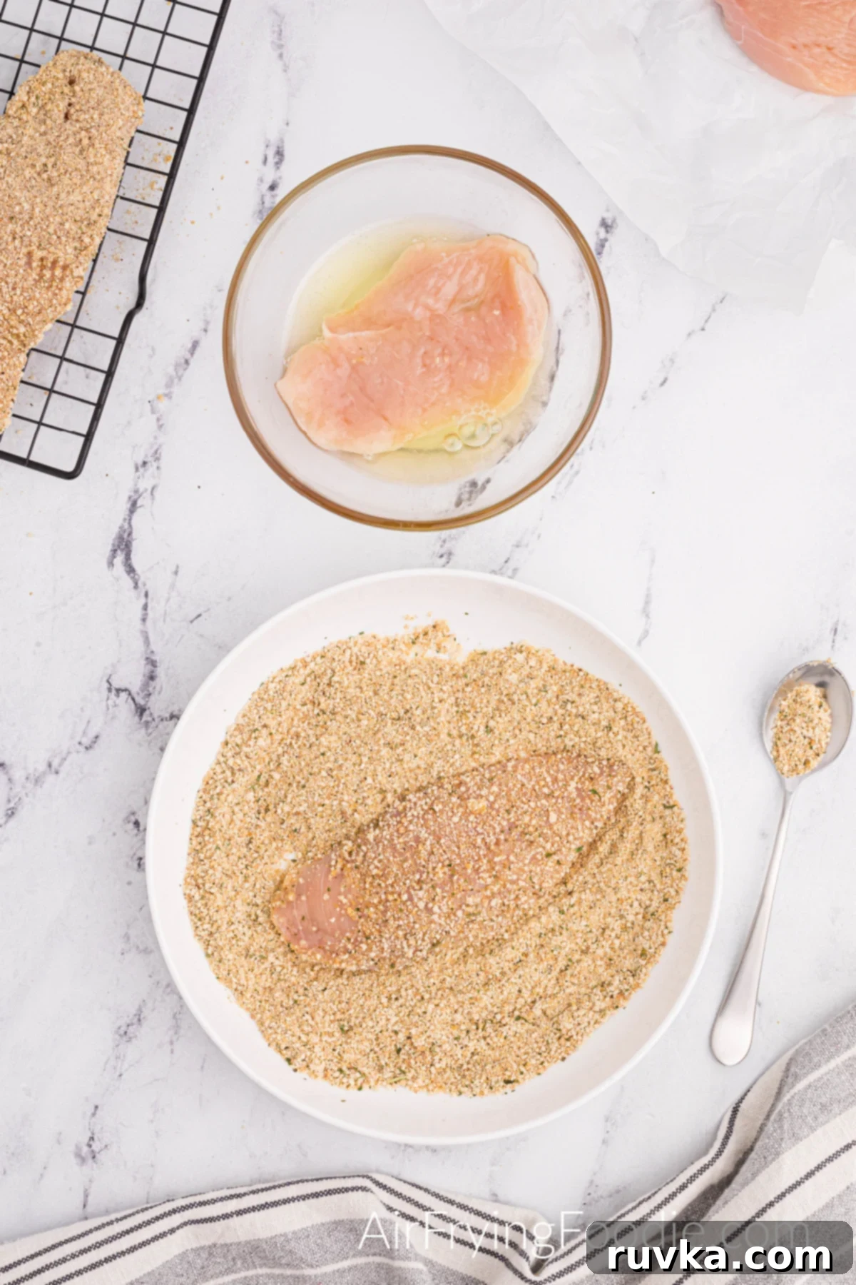 Perfectly Golden Air Fryer Chicken Cutlets 4 A round white bowl with breadcrumb mixture, a chicken cutlet is resting inside the bowl covered with breadcrumbs, and a small silver spoon of breadcrumbs. A second round clear bowl with egg whites and a chicken cutlet covered with egg whites and prepared to go in the breadcrumb mixture.