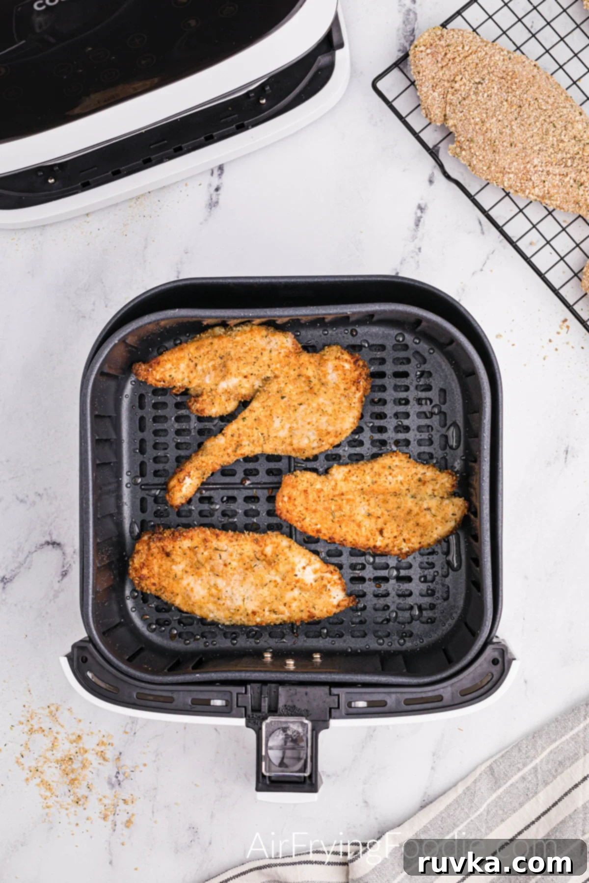 Perfectly Golden Air Fryer Chicken Cutlets 6 Three cooked chicken cutlets in the air fryer basket with a small portion of the air fryer base and wire cooling rack with uncooked chicken cutlets at the top of the picture.