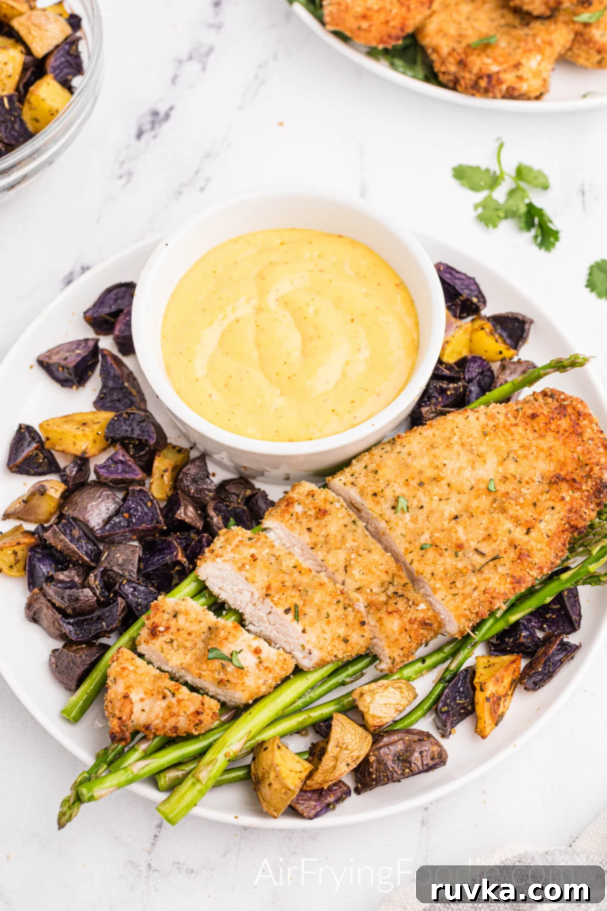 Perfectly Golden Air Fryer Chicken Cutlets 7 Overhead picture of air fryer chicken cutlets fully cooked on a round white plate with half the chicken cut up into strips served with asparagus, fingerling potatoes, and a bowl of creamy honey mustard sauce. At the top of the picture is a small portion of a glass bowl with extra fingerling potatoes and a second round plate with a stack of fully cooked chicken cutlets.