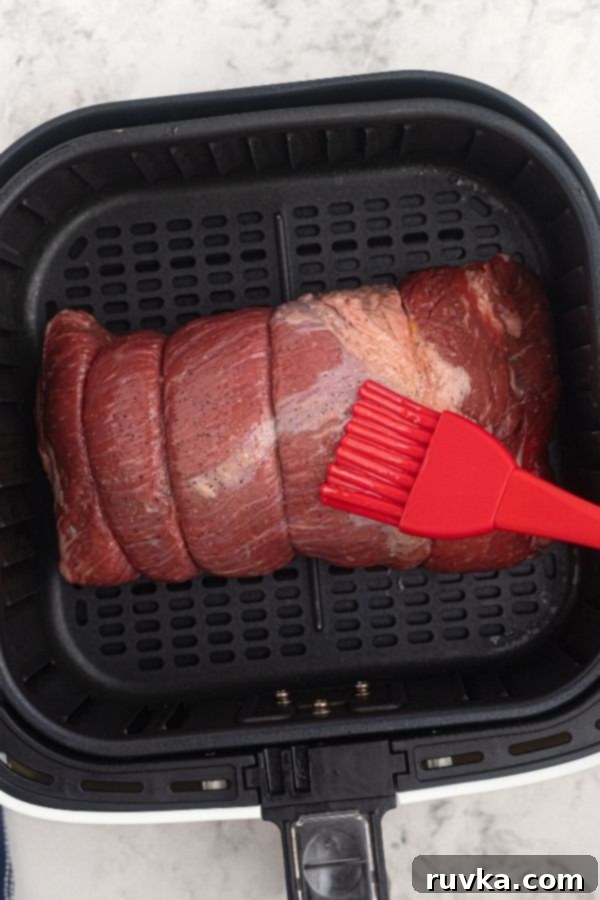 Crispy Air Fryer Top Round 3 Uncooked top round roast in the air fryer basket, being brushed with olive oil, and seasoned with salt and pepper.