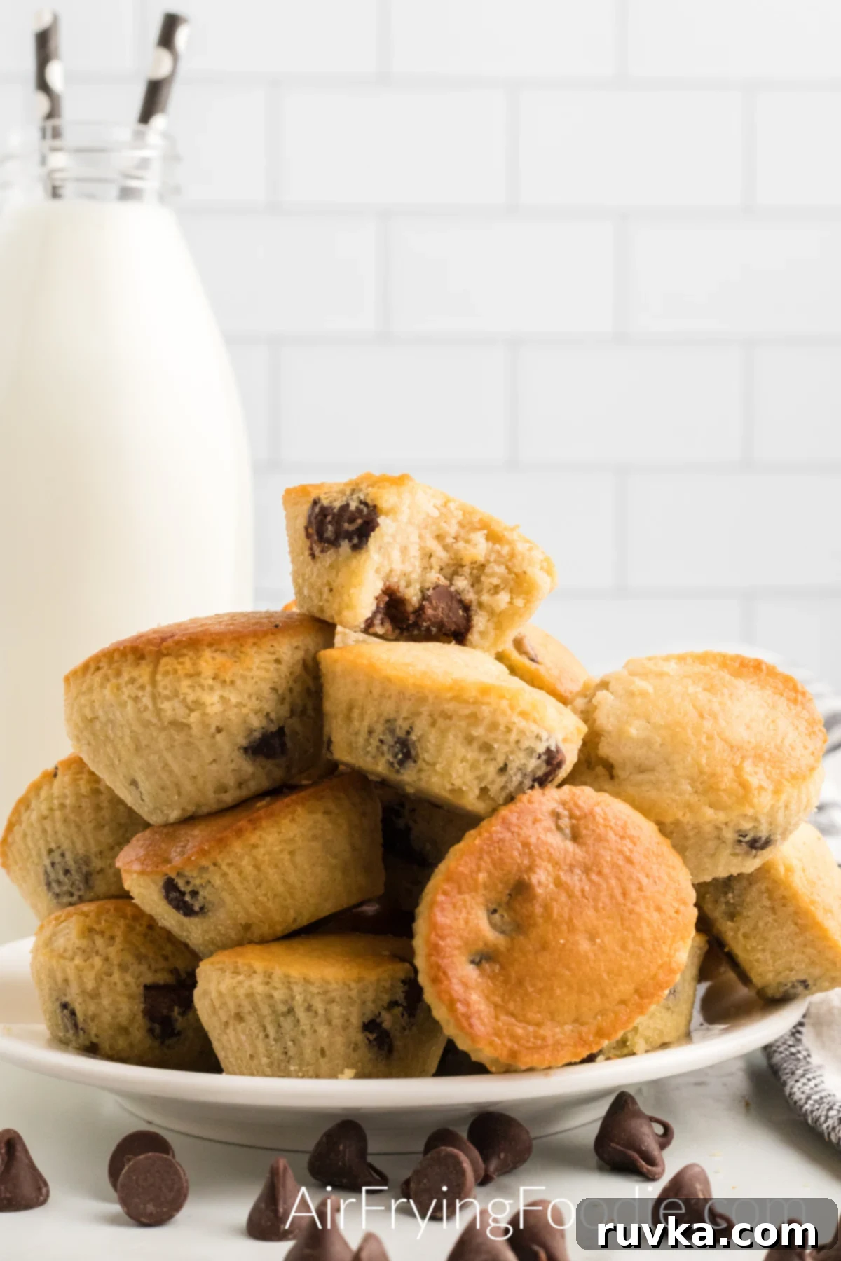 Easy Air Fryer Chocolate Chip Muffins 2 Golden-brown chocolate chip muffins, freshly cooked in an air fryer, stacked on a pristine white plate. Scattered chocolate chips rest on the table in front, accompanied by an elegant tall glass bottle of milk with two straws, hinting at the perfect pairing.