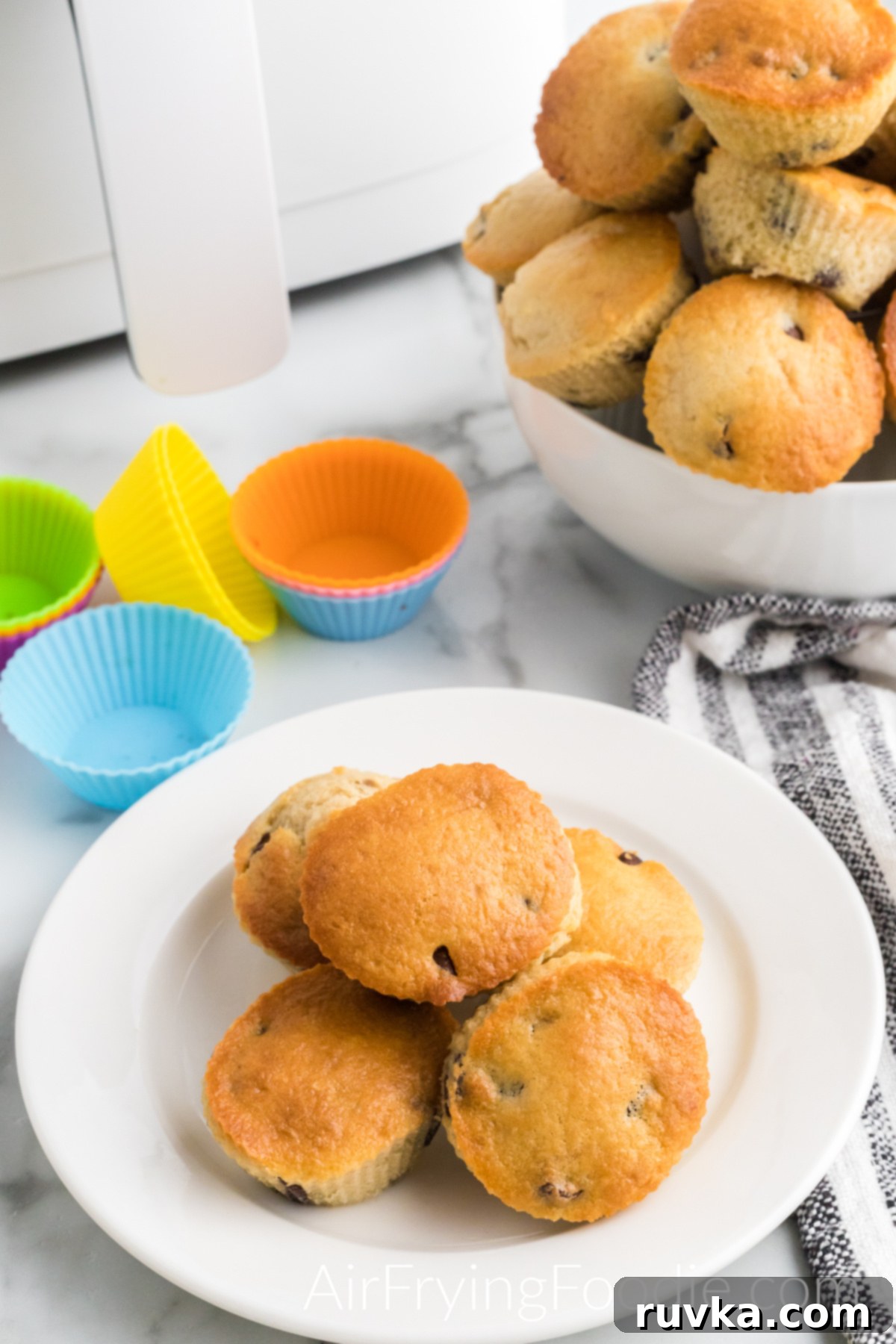 Easy Air Fryer Chocolate Chip Muffins 9 A delightful arrangement of freshly baked air fryer chocolate chip muffins. Five golden-brown muffins are artfully served on a small, elegant round white plate, while a generous bowl overflowing with more muffins sits in the top right. Above the serving plate, several colorful silicone muffin cups are neatly stacked, and the air fryer base is subtly visible in the top right, a testament to the easy baking process.
