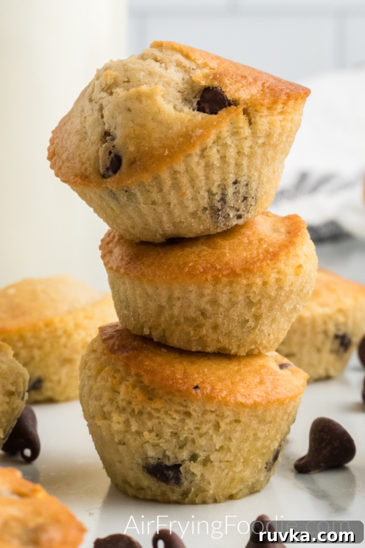 Easy Air Fryer Chocolate Chip Muffins 10 A charming stack of three golden-brown air fryer chocolate chip muffins, perfectly baked and inviting. More delicious muffins are visible beside and behind the stack, creating a tempting display. A scattering of rich chocolate chips on the table in front of the stack adds a final touch of sweetness.