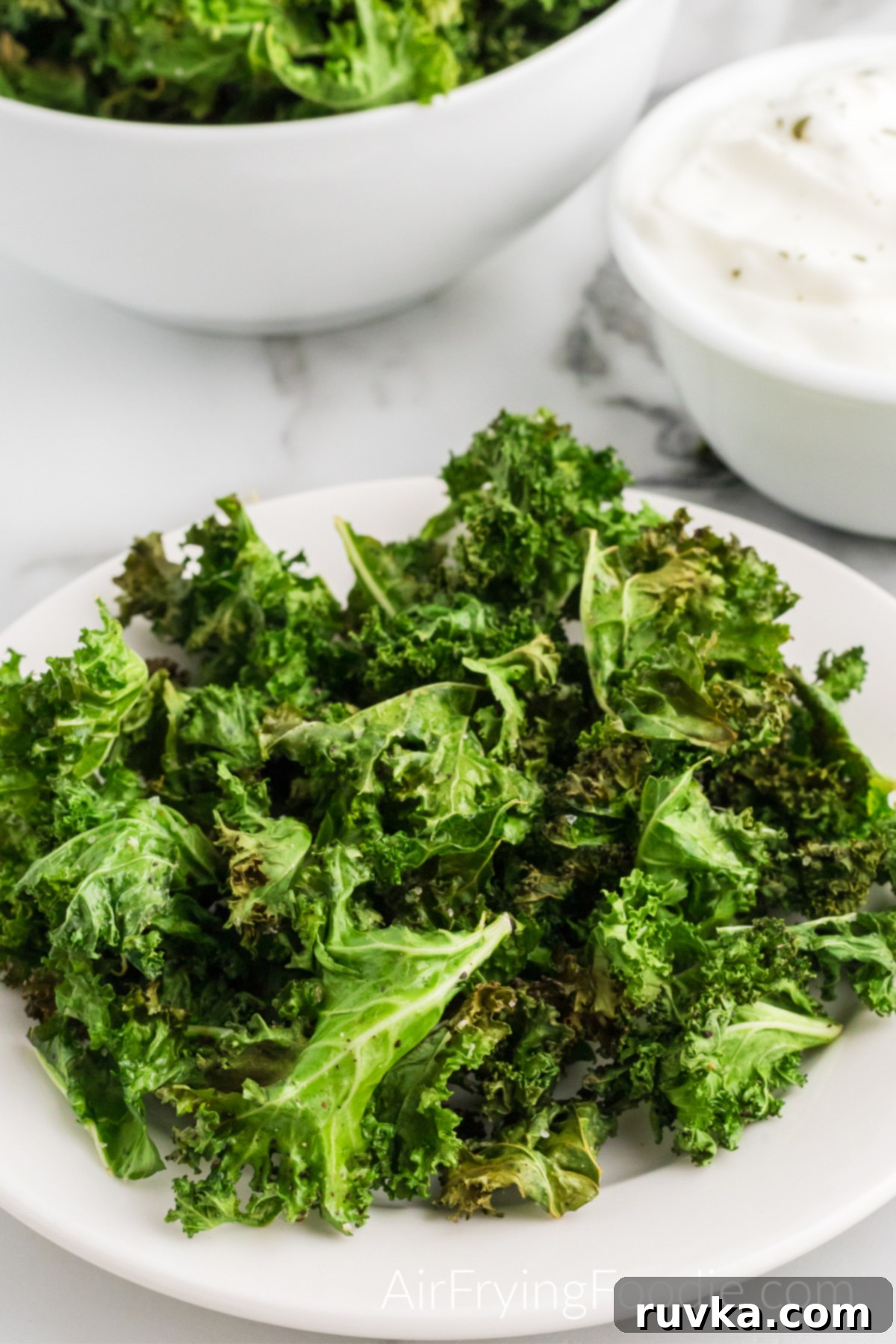Crispy Air Fryer Kale Chips 2 A round white plate of kale chips cooked in the air fryer. In the top left corner of the picture is a round white bowl with extra kale chips and a greek yogurt dipping sauce in a white bowl in the top right corner.