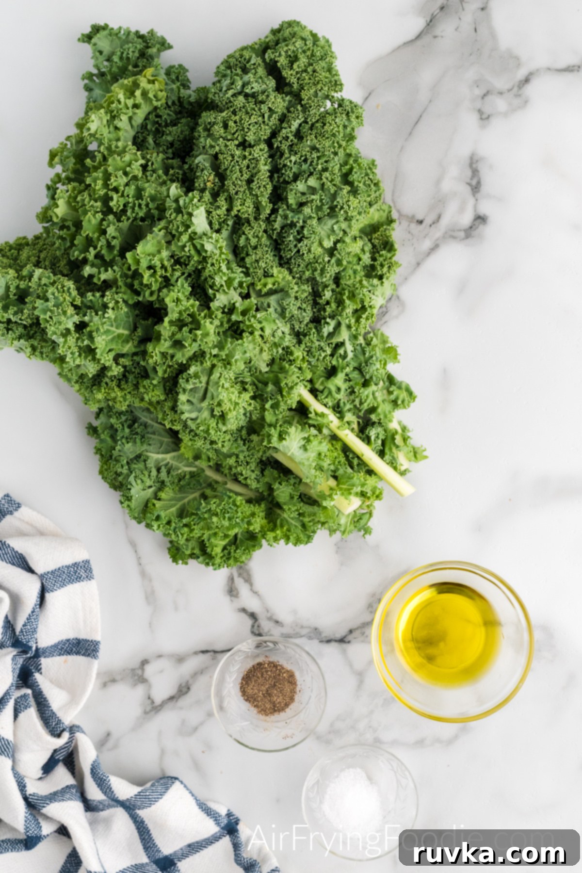 Crispy Air Fryer Kale Chips 3 Ingredients picture with everything you will need to make kale chips in the air fryer. At the bottom right of the image are three small glass bowls, each containing a different ingredient; sea salt, black pepper, and olive oil. There is a large bundle of kale above the glass bowls.