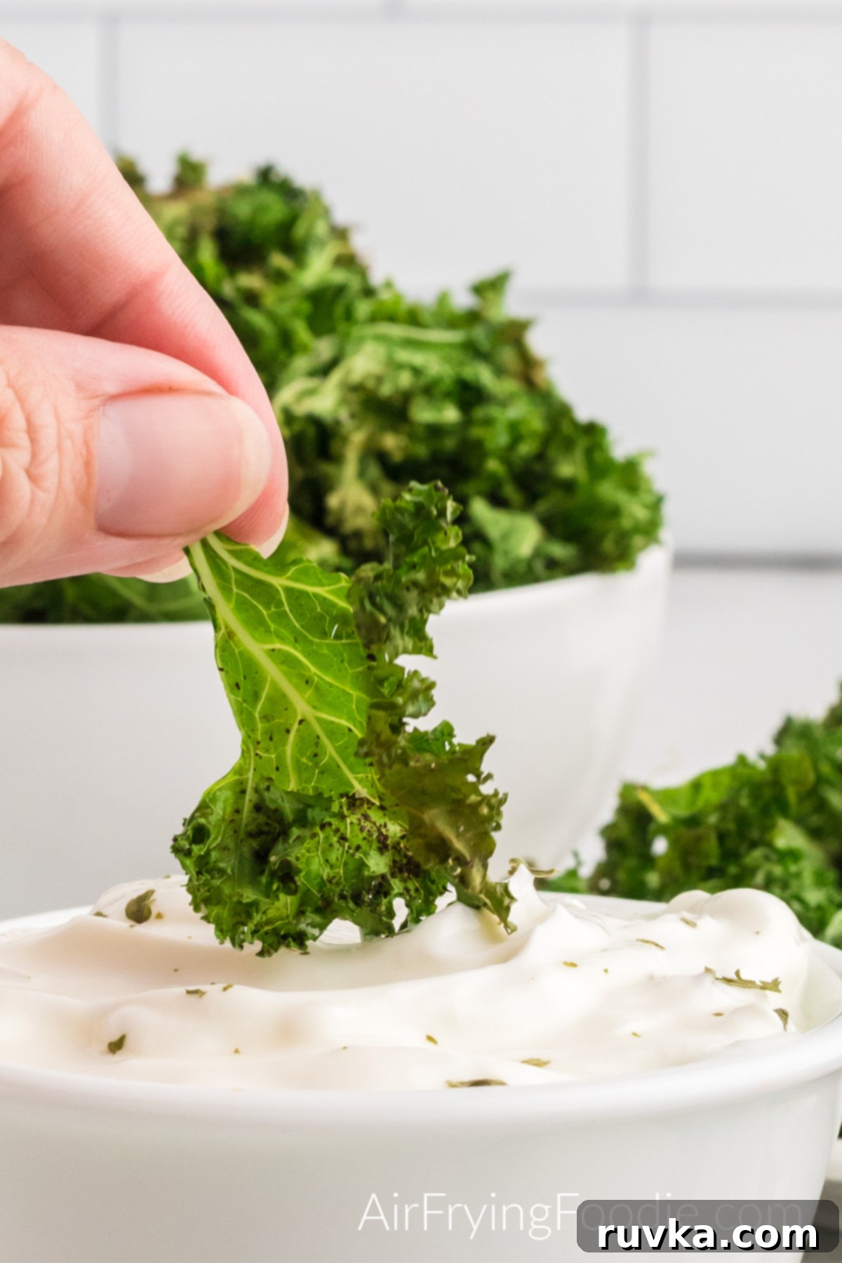 Crispy Air Fryer Kale Chips 9 Close-up picture of kale chips cooked in the air fryer being dipped in greek yogurt. Behind the bowl of greek yogurt is a white bowl of kale chips.