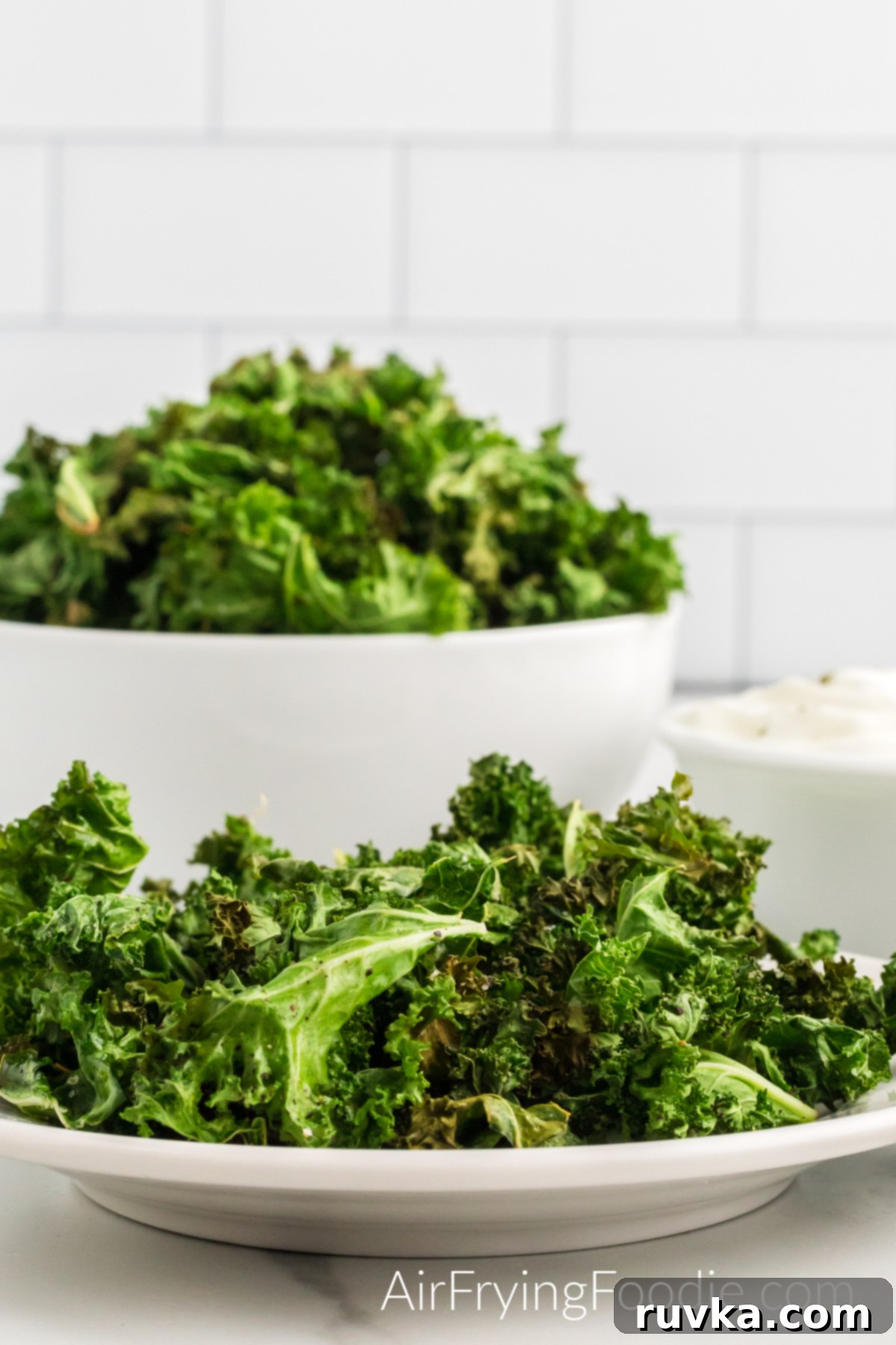 Crispy Air Fryer Kale Chips 10 A close-up picture of kale chips cooked in the air fryer served on a small round white plate. Behind the small plate is a round bowl with more kale chips and a small round white bowl of yogurt dip to the right of the bowl.