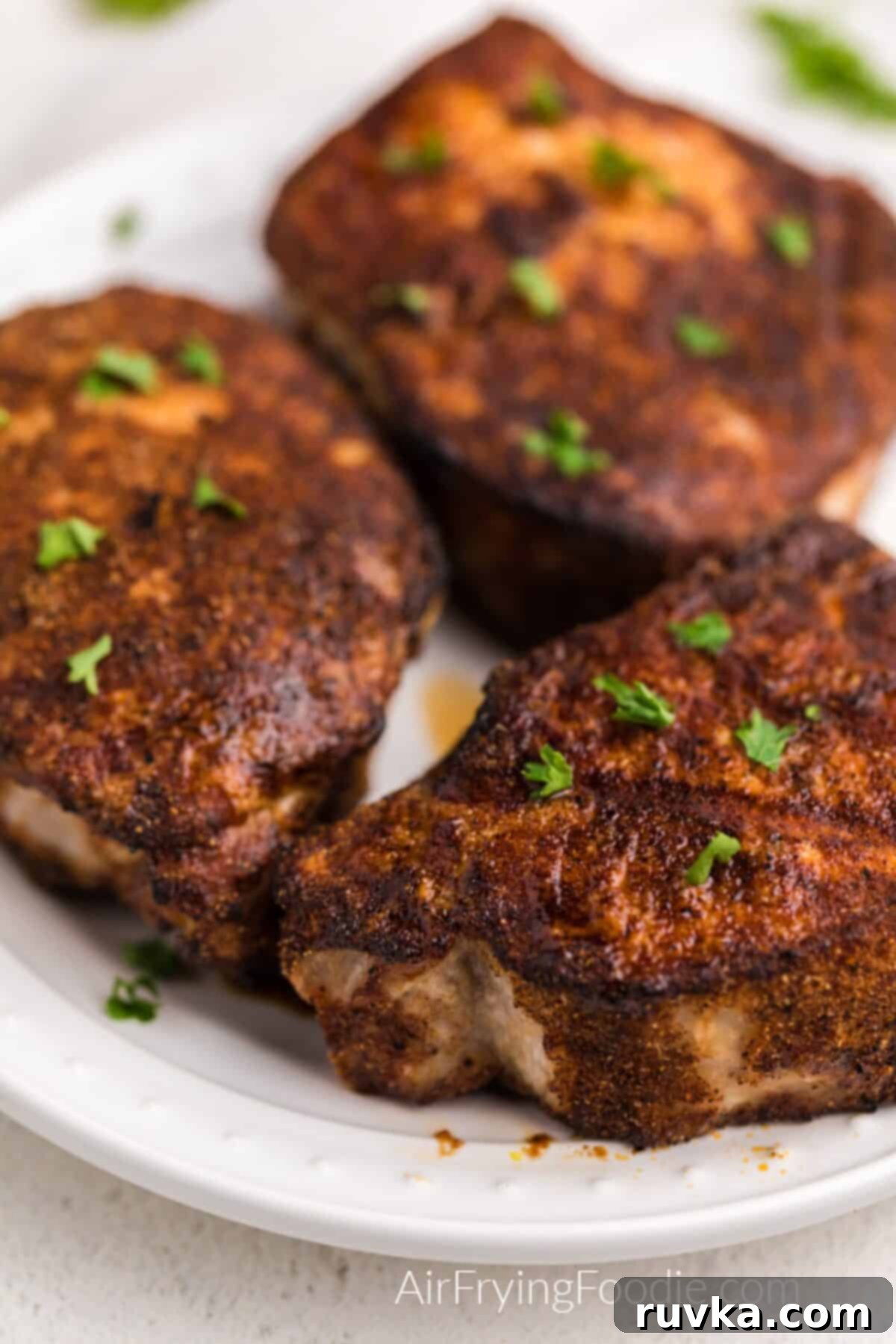 Juicy Air Fryer Pork Chops 3 Close up photo of thick cut seasoned and air-fried pork chops on a white plate, ready to be served. The chops have a rich, golden brown color.