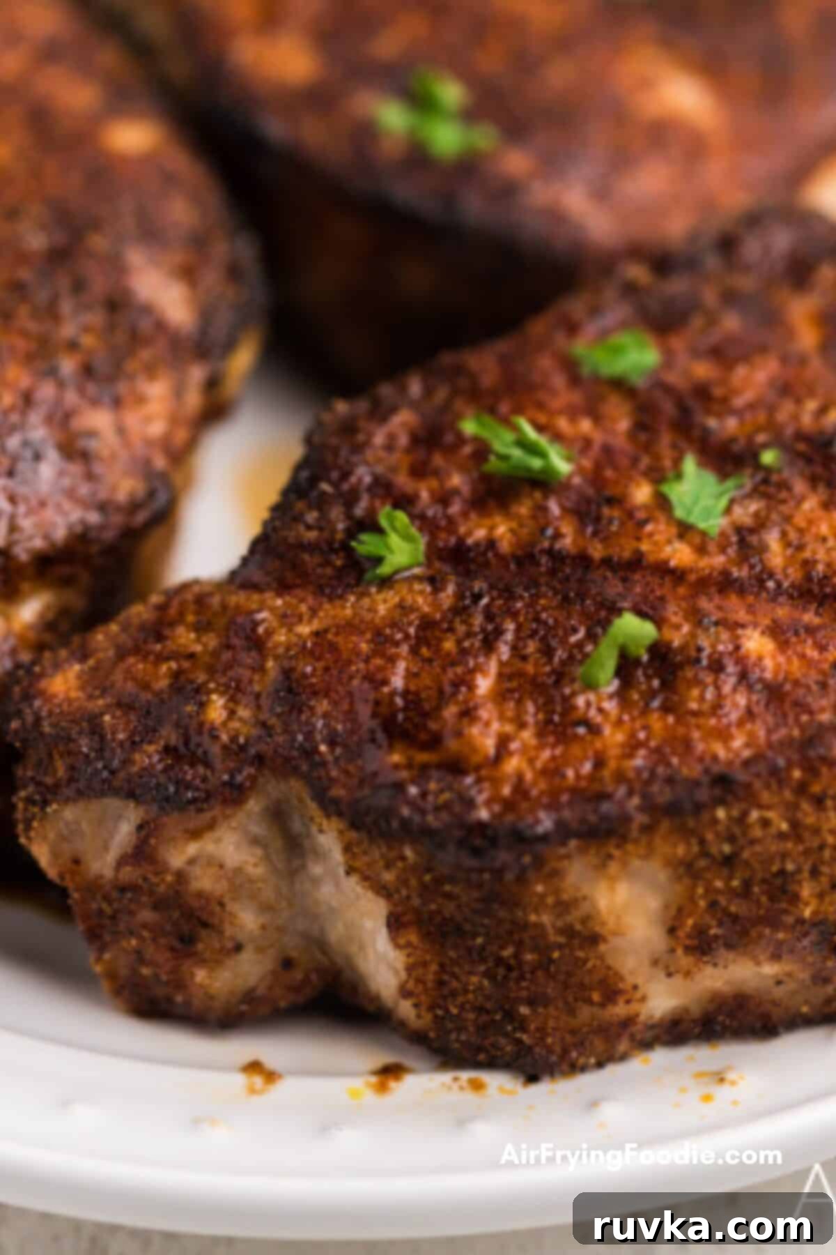 Juicy Air Fryer Pork Chops 4 Crispy, seasoned, air fryer pork chops on a white plate, ready to eat. They are garnished with fresh herbs and look perfectly cooked.