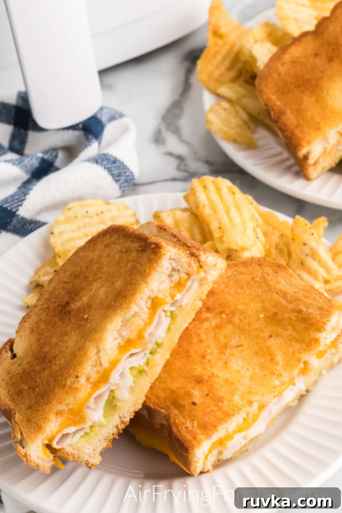 Crispy Air Fryer Turkey Cheese Melt 2 Air fryer turkey cheese melt cut in half with melted cheese, fresh avocado, and a side of ruffle chips served on a small round plate. A second small round plate with turkey cheese melt and chips is in the top right of the picture, and the base of the air fryer is partially showing in the top left of the image.