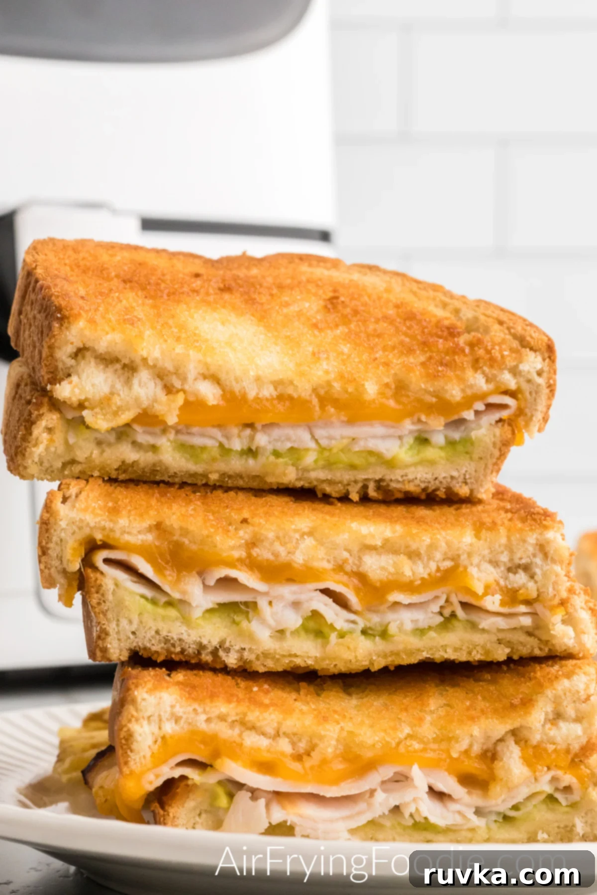 Crispy Air Fryer Turkey Cheese Melt 11 Three turnkey cheese melts are cut in half and stacked on a round white plate after being cooked in the air fryer. You can see the melted cheese, turkey, and avocado. The base of the air fryer is in the top left of the image.