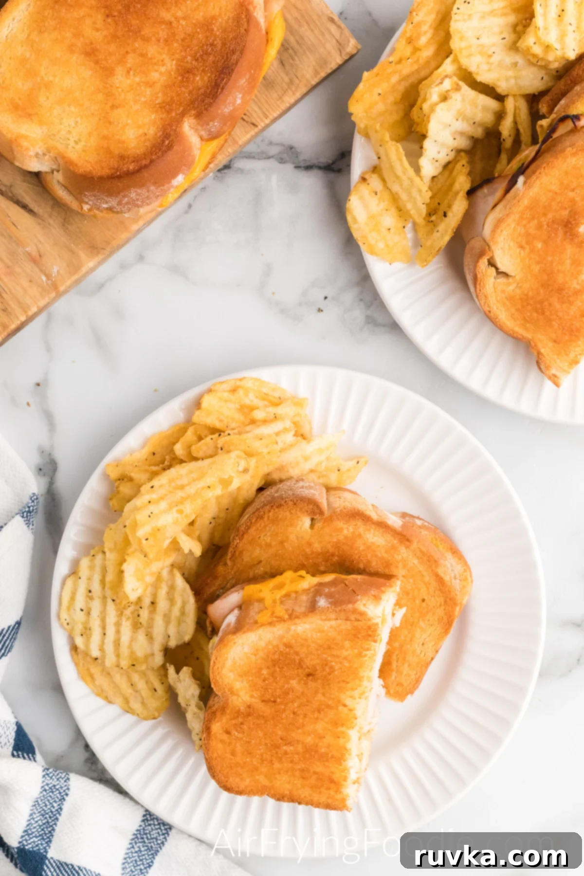 Crispy Air Fryer Turkey Cheese Melt 12 Overhead shot of a turkey cheese melt cooked in the air fryer, cut in half, and served with a side of potato chips on a round white plate. There is a small portion of a second turkey cheese melt on a round white plate in the top right of the picture and an extra sandwich on a cutting board in the top left of the image.