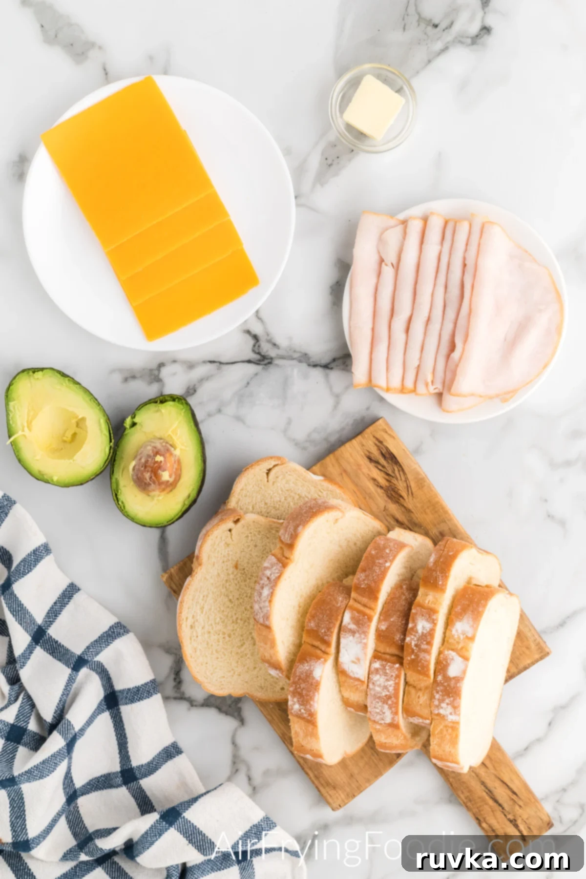 Crispy Air Fryer Turkey Cheese Melt 3 Ingredients picture everything you need to make your air fryer turkey cheese melt. Starting at the bottom of the picture to the top is a cutting board with eight slices of white bread, one avocado cut in half, two small round white plates, one plate with turkey and the second plate with cheese, and a small round glass bowl with butter.