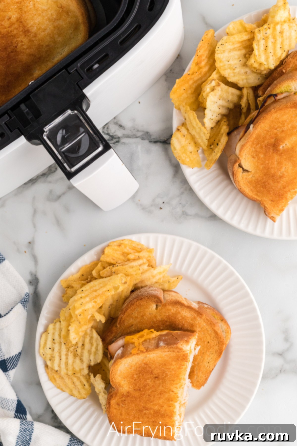 Crispy Air Fryer Turkey Cheese Melt 10 Overhead picture of an air fryer turkey cheese melt, cut in half and served with a side of ruffle potato chips on a round white plate. There is a second round white plate with another turkey cheese melt and chips in the top right of the picture, and a small portion of the air fryer base is in the top left.