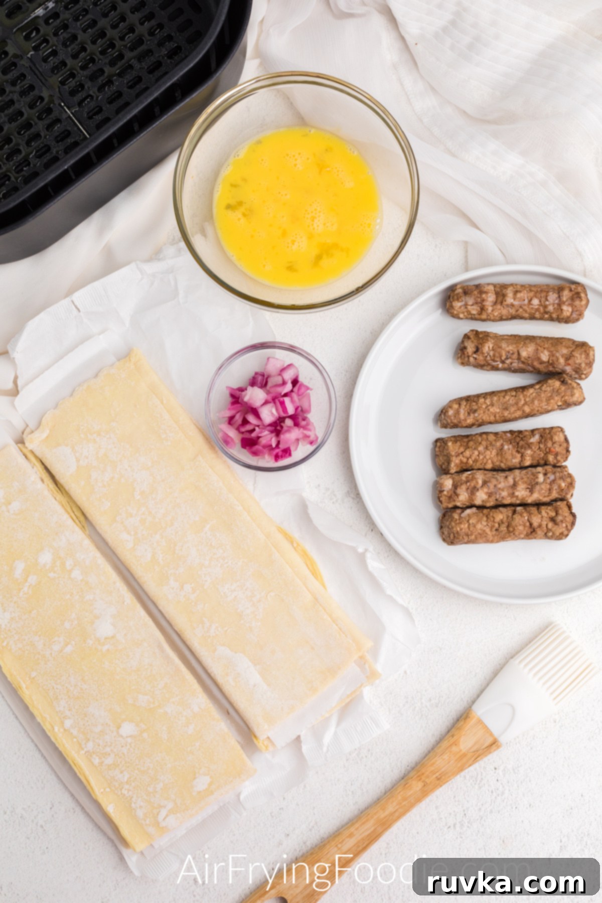 Perfect Air Fryer Sausage Rolls 3 puff pastry, diced onion, beaten egg, and sausage links on a table ready to be made into sausage rolls. A neat setup for efficient cooking.
