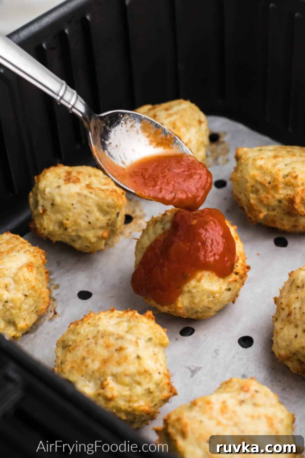Crispy Air Fryer Chicken Parmesan Meatballs 4 Chicken meatballs in air fryer basket on top of parchment paper, being topped with marinara sauce.