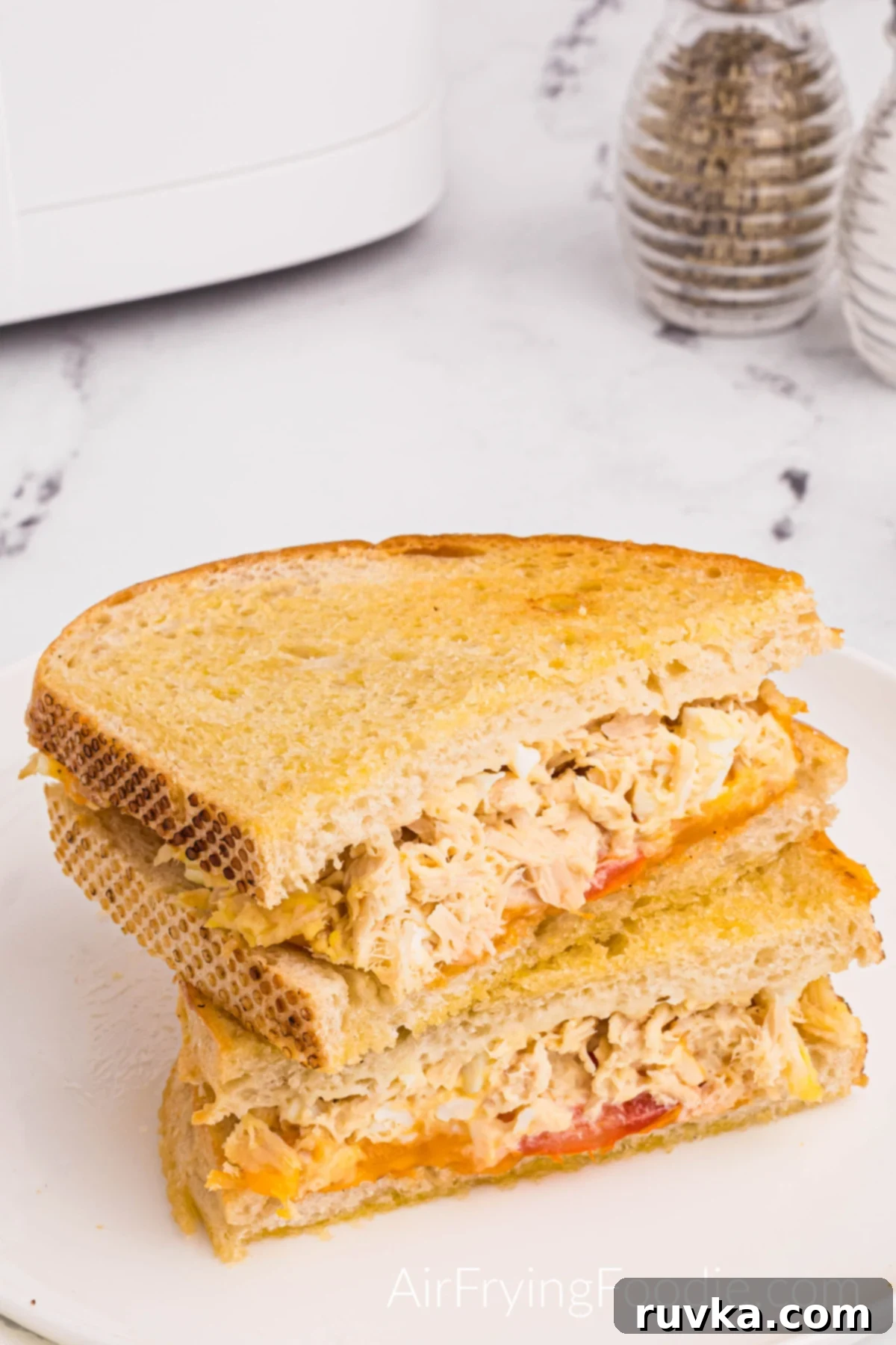 Crispy Air Fryer Tuna Melt 2 Tuna melt on golden Texas toast cooked in the air fryer with tomato and cheese cut in half and served on a round white plate. A small portion of the air fryer base is in the top left of the picture, and a sale and pepper shaker is in the top right.
