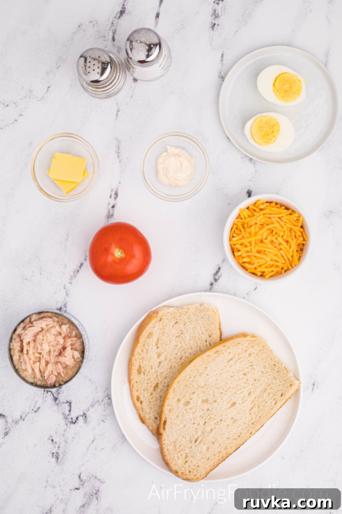 Crispy Air Fryer Tuna Melt 3 Overhead shot of the ingredients used to make a tuna melt in the air fryer. Two round white plates; one with two slices of Texas toast and the second with one egg boiled and cut in half. Three small glass bowls contain tuna; the other has butter and mayonnaise—a small white bowl of shredded cheese, one whole tomato, and a salt and pepper shaker.