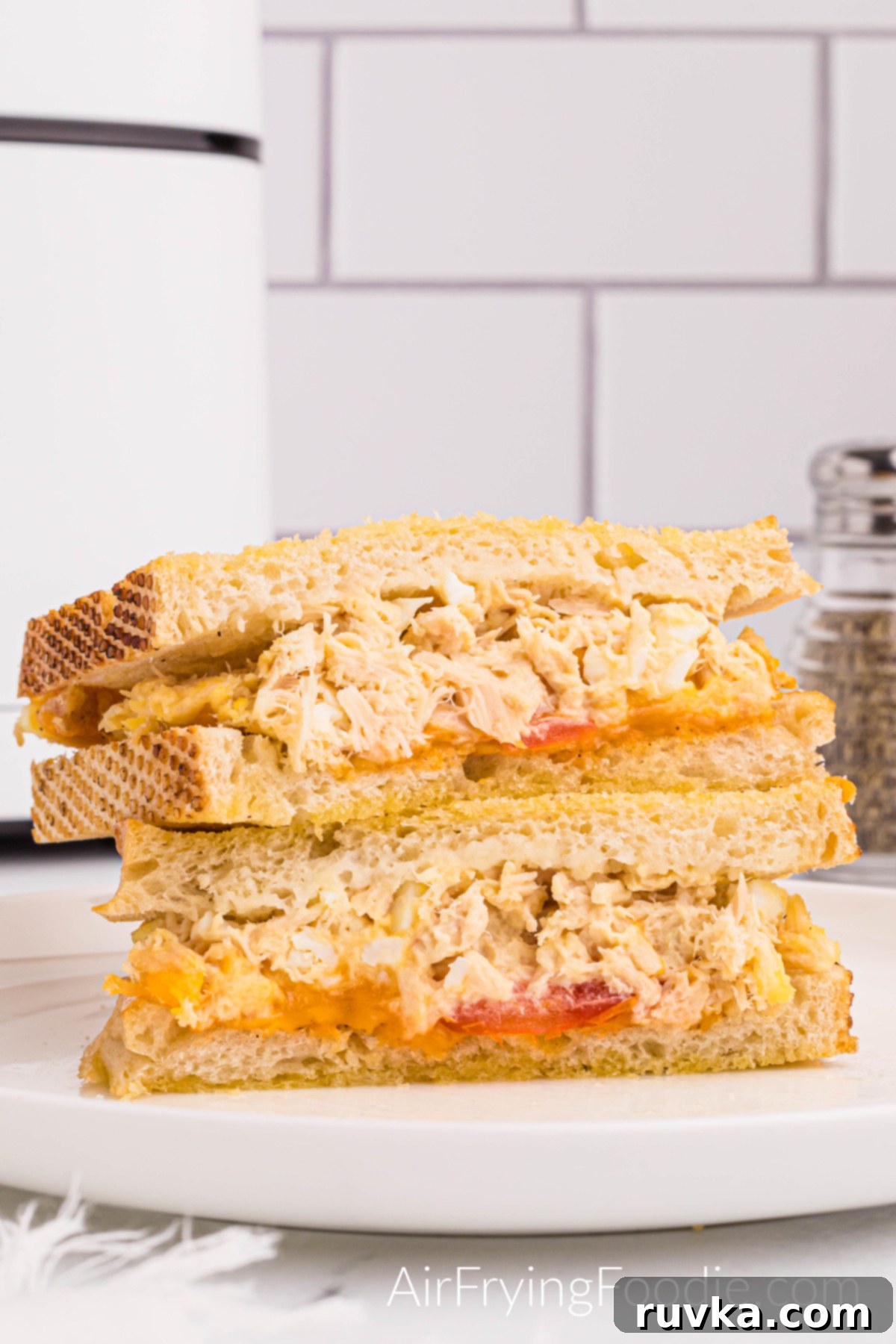 Crispy Air Fryer Tuna Melt 10 Tuna melt sandwich on golden Texas toast cooked in the air fryer with tomato and cheese cut in half and served on a round white plate. There is a small portion of a pepper shaker to the right of the sandwich and a small part of the air fryer base to the left of the picture.