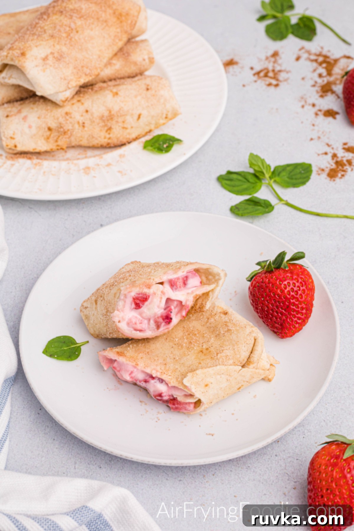 Crispy Air Fryer Strawberry Cheesecake Chimis 2 Air fryer strawberry cheesecake chimichanga cut in half, partially stacked and served on a round white plate with a whole strawberry and basil leaf as a garnish. A portion of a whole strawberry is in the bottom right corner of the picture. There is a round white plate in the top left of the photograph stacked with more strawberry chimichangas, and to the right is more basil garnish and a small portion of a strawberry showing.