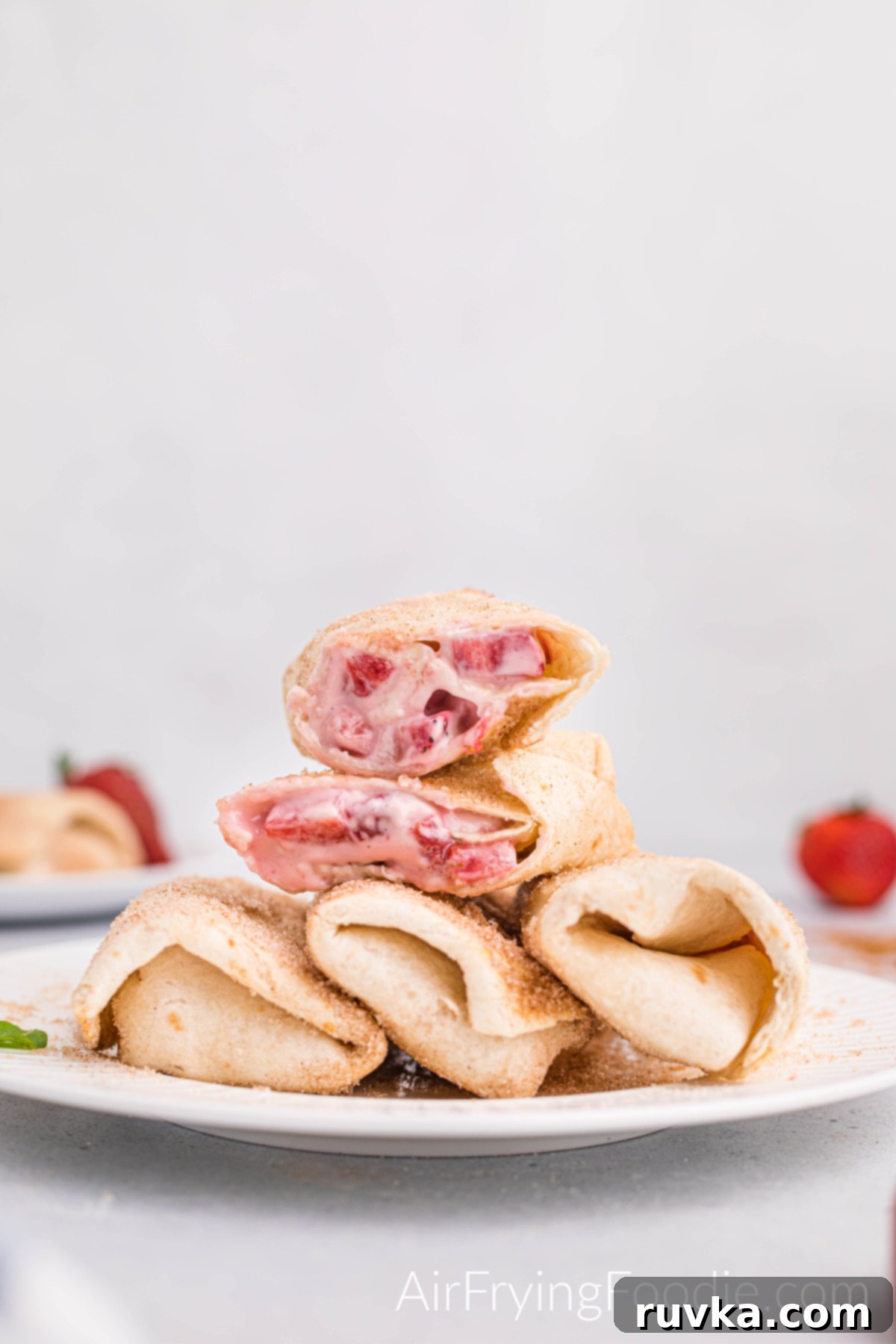 Crispy Air Fryer Strawberry Cheesecake Chimis 13 Close-up picture of air fryer strawberry cheesecake chimichangas served on a white plate. Three are lined on the plate, and one is cut in half and stacked on top of the three. In the background is a blurred image of another plate of chimichangas and to the right is a blurred image of a whole strawberry.