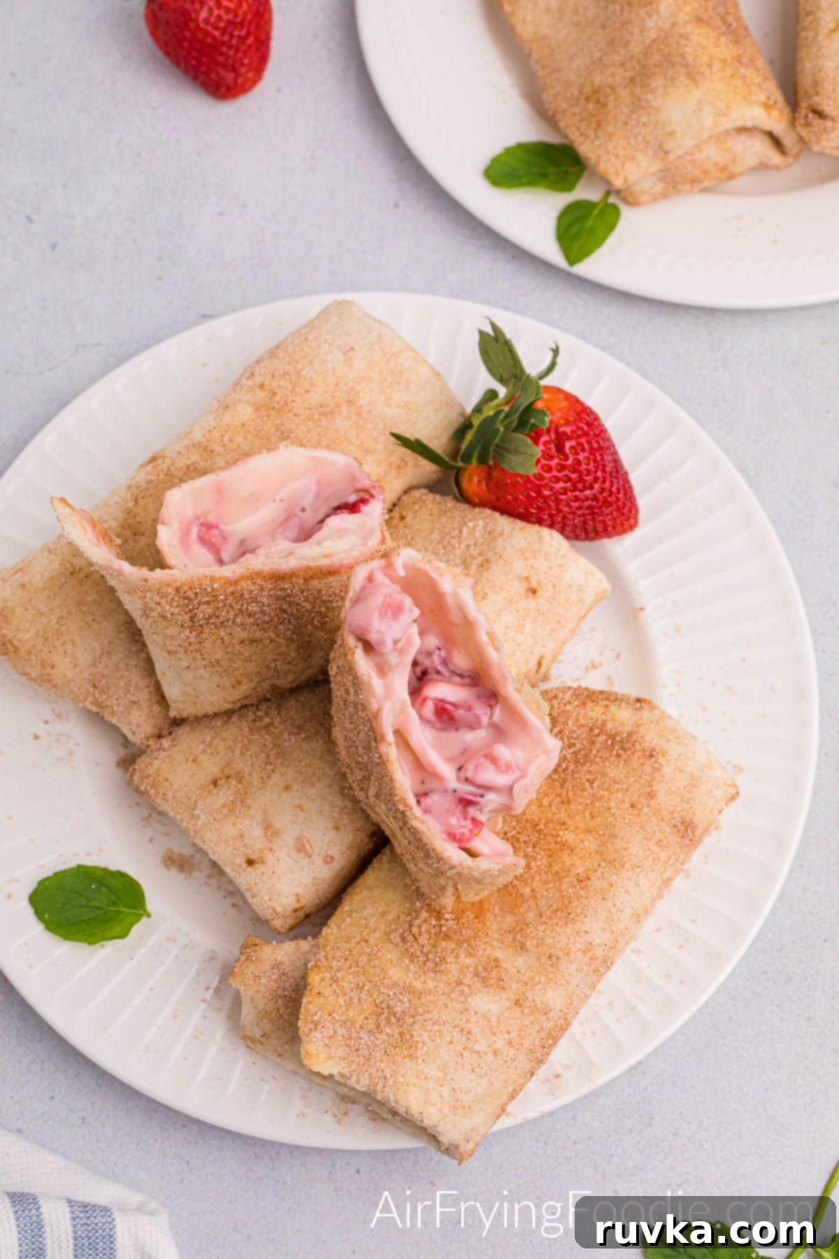 Crispy Air Fryer Strawberry Cheesecake Chimis 14 Overhead shot of three strawberry cheesecake chimichangas cooked in the air fryer and lined on a round white plate. A fourth chimichanga is cut in half, stands on top of the three chimichangas, and is garnished with a whole strawberry. In the top right of the picture is another round white plate with two chimichangas partially showing, and a whole strawberry is to the left.